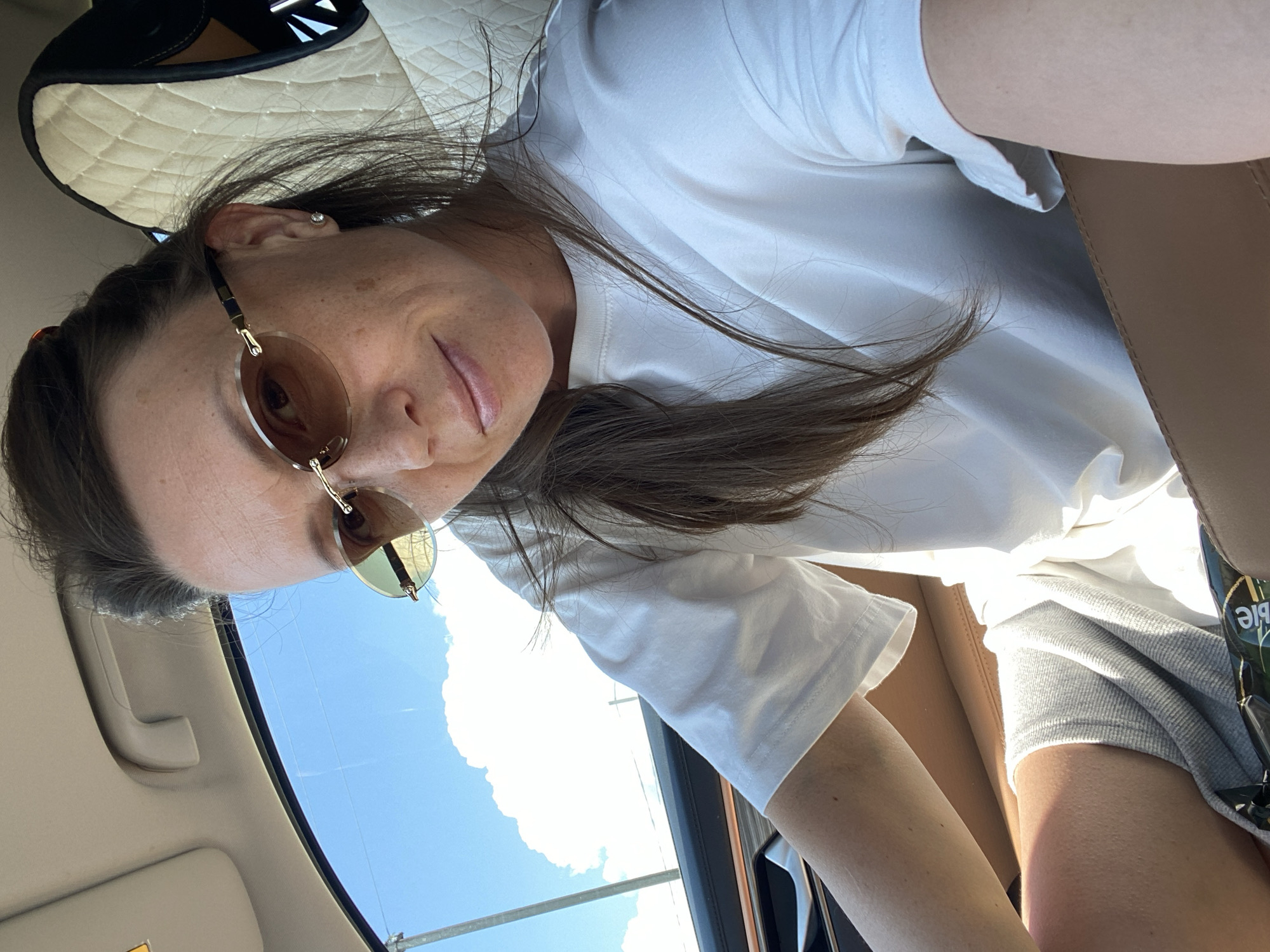 A woman is sitting in the driver's seat of a car. She is wearing sunglasses and a white t-shirt. The car's interior is visible, with a tan leather seat and quilted headrest. Outside the car, the sky is blue with a large white cloud. The location is identified as Kvards, Russia, which is a village in the Kurchaloyevsky District of the Chechen Republic, Russia.
