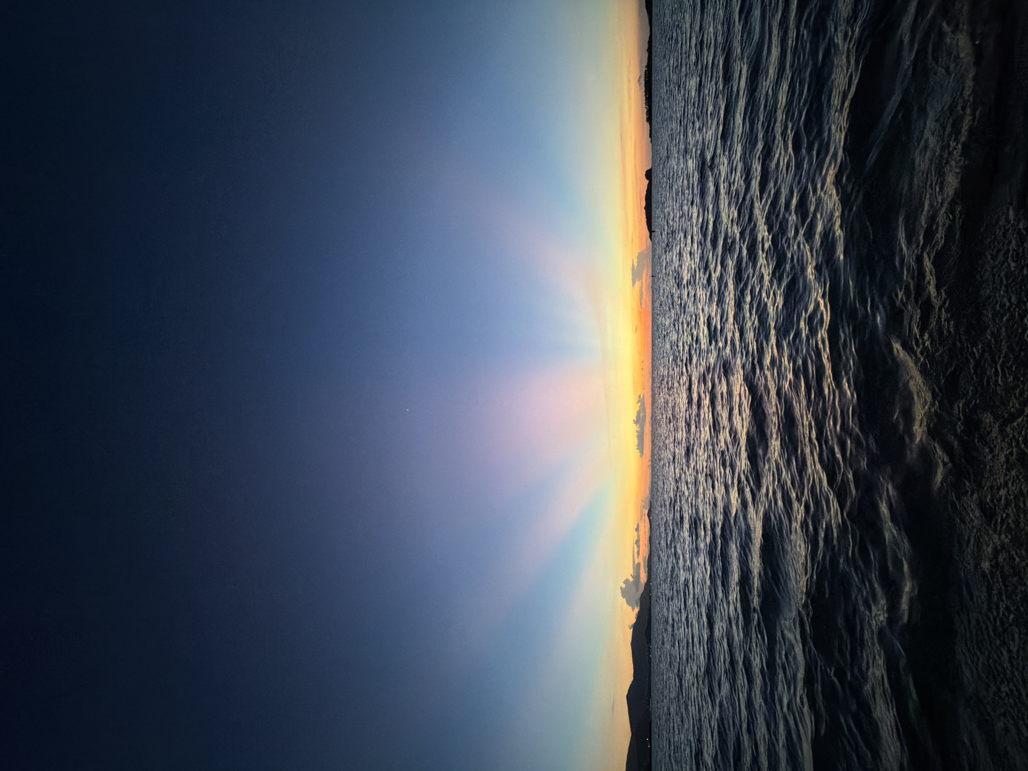 This image depicts a serene seascape at sunset. The scene is divided horizontally, with the left side showcasing a gradient of the sky transitioning from a deep, dark blue to lighter shades of purple, pink, and yellow as it approaches the horizon. Rays of light emanate from the sun, which is just below the horizon, creating a dramatic visual effect.
The right side of the image features the ocean, with choppy waves creating a textured surface. The water reflects the warm colors of the sunset, with streaks of gold and orange visible amidst the darker blues and grays of the waves. In the far distance, a silhouette of landmass, likely mountains or hills, is visible along the horizon. The overall mood is peaceful and awe-inspiring, characteristic of a beautiful sunset. The time of day is clearly sunset. There are no visible people or objects in the image, nor any visible text. The location context suggests this is Baan Plai Laem, Thailand.