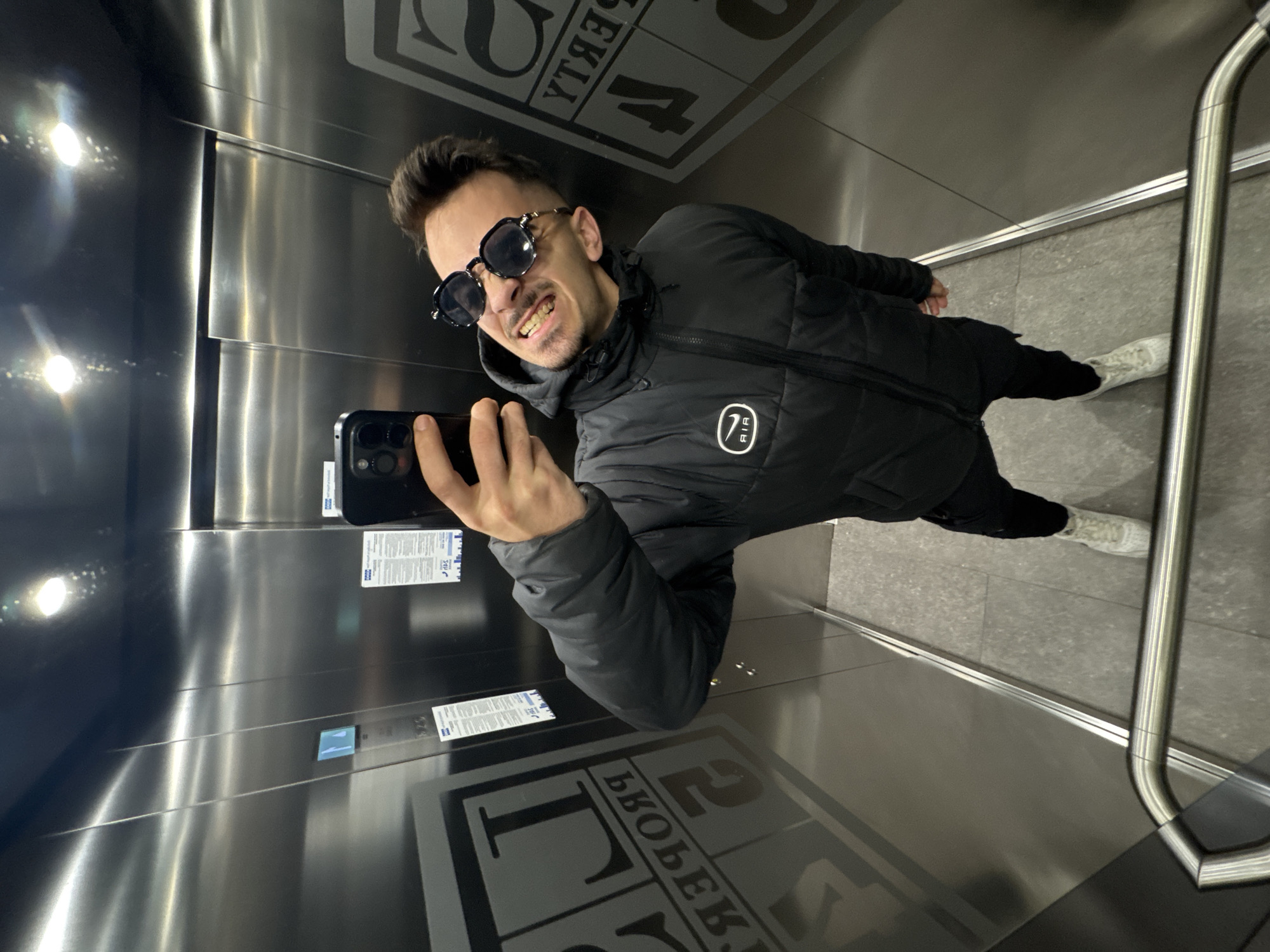 A man is taking a selfie in an elevator. The elevator has stainless steel walls and a metal handrail. Floor tiles are visible and have a dark grey pattern on them. Stickers are affixed to the walls, in Cyrillic script, implying a location in Bulgaria. The reflection of interior lights is visible on the walls. The man is wearing a black puffer jacket, dark pants, and white shoes. He is also wearing sunglasses. A "7" logo is visible on the jacket. The city is Sofia, Bulgaria.