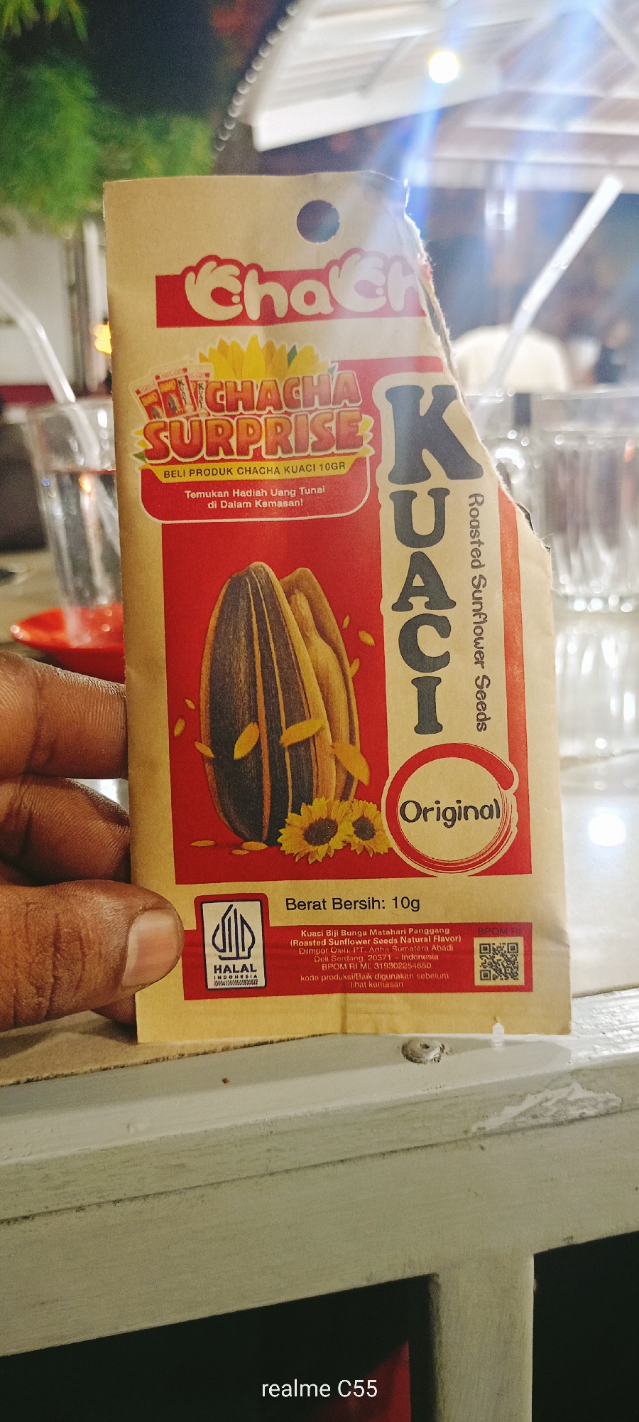 The image shows a package of "Cha Cha Kuaci" roasted sunflower seeds, being held by a dark-skinned hand with a visible fingerprint. The package is made of brown paper with red and white printing. It features the brand name "Cha Cha," the word "Kuaci" (Indonesian for sunflower seeds), an image of sunflower seeds, and the word "Original." The text also mentions a "Surprise" and a promotion involving finding cash prizes inside the package. Information about the product's weight (10g), ingredients, and Halal certification are visible. A QR code is also present. The background is blurry, but hints at a table setting with glasses and a red object. The text "realme C55" is at the bottom. It is not possible to determine the location Lhokseumawe, Indonesia from the image.