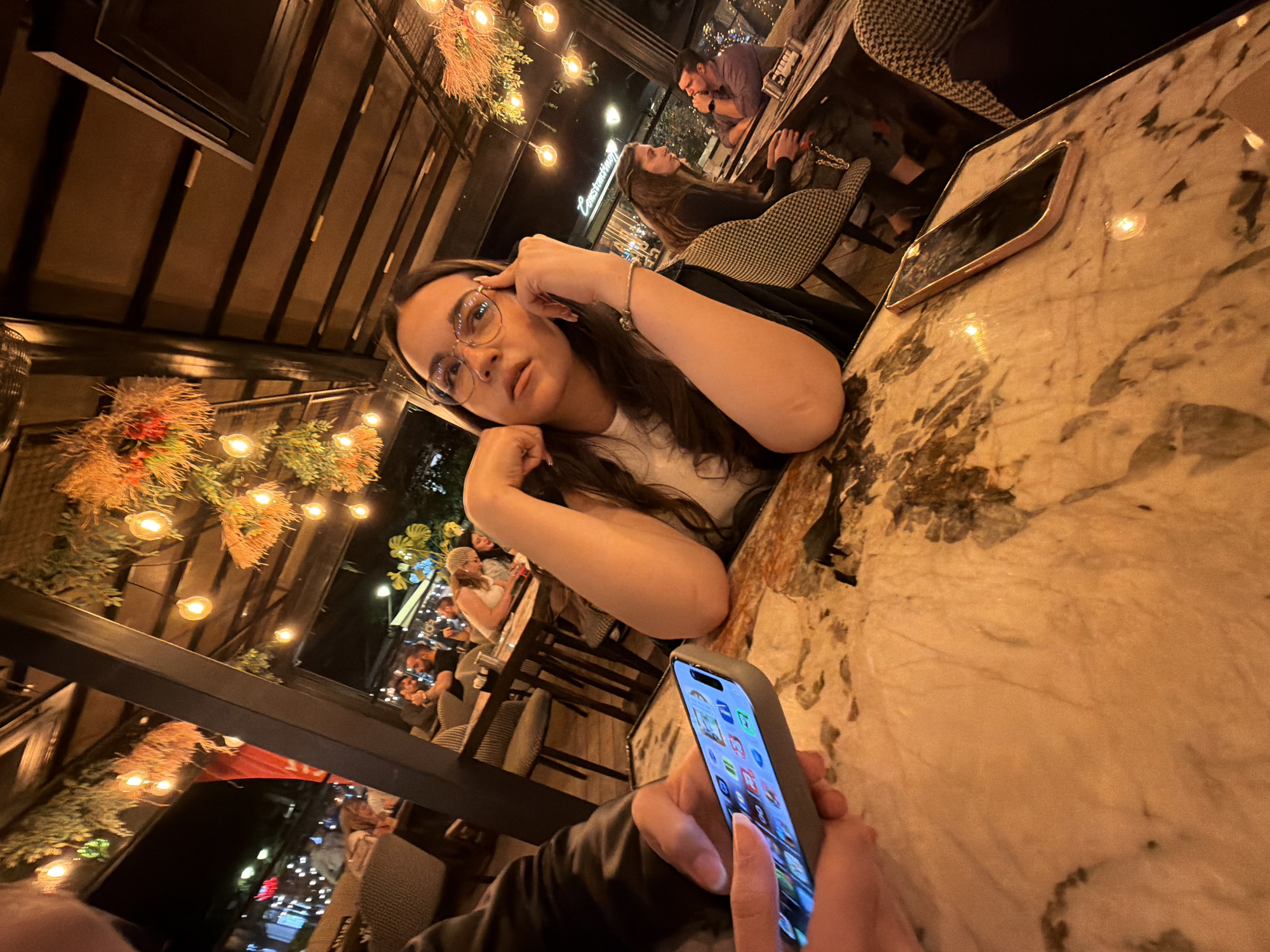 A person holds a smartphone while seated at a table with a woman. The woman is wearing glasses and has her head resting on her hand. The table is marble and has another smartphone on it. The setting appears to be an outdoor restaurant or patio, with other people in the background and decorative lights overhead. The location is in Sofia, Bulgaria, indicated by a sign "Constantinesti" visible in the background.
