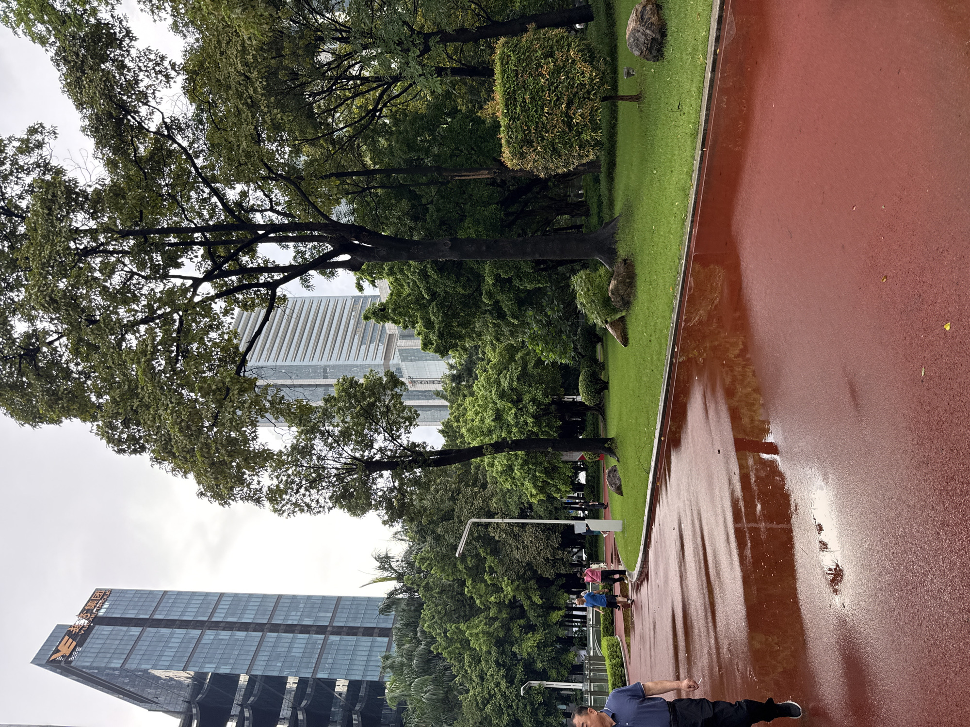 The image captures a scene in a park in Tianhe District, China, on a cloudy day. In the foreground, a red running track is wet and reflects the surroundings, with puddles scattered across its surface. A man in a dark blue polo shirt and black pants is walking on the track from the left side of the frame. In the midground, several people are walking on a paved path behind the running track. To the right, a lush green lawn is dotted with rocks, a neatly trimmed bush, and trees.
Dominating the background are modern skyscrapers, indicative of an urban environment. A tall, glass-fronted building with a prominent logo at the top, displaying Chinese characters and "YUEXIU GROUP," is visible on the left. Another tall building with horizontal striped panels stands further back. The sky is overcast and gray. The overall impression is a blend of urban development and green public space.