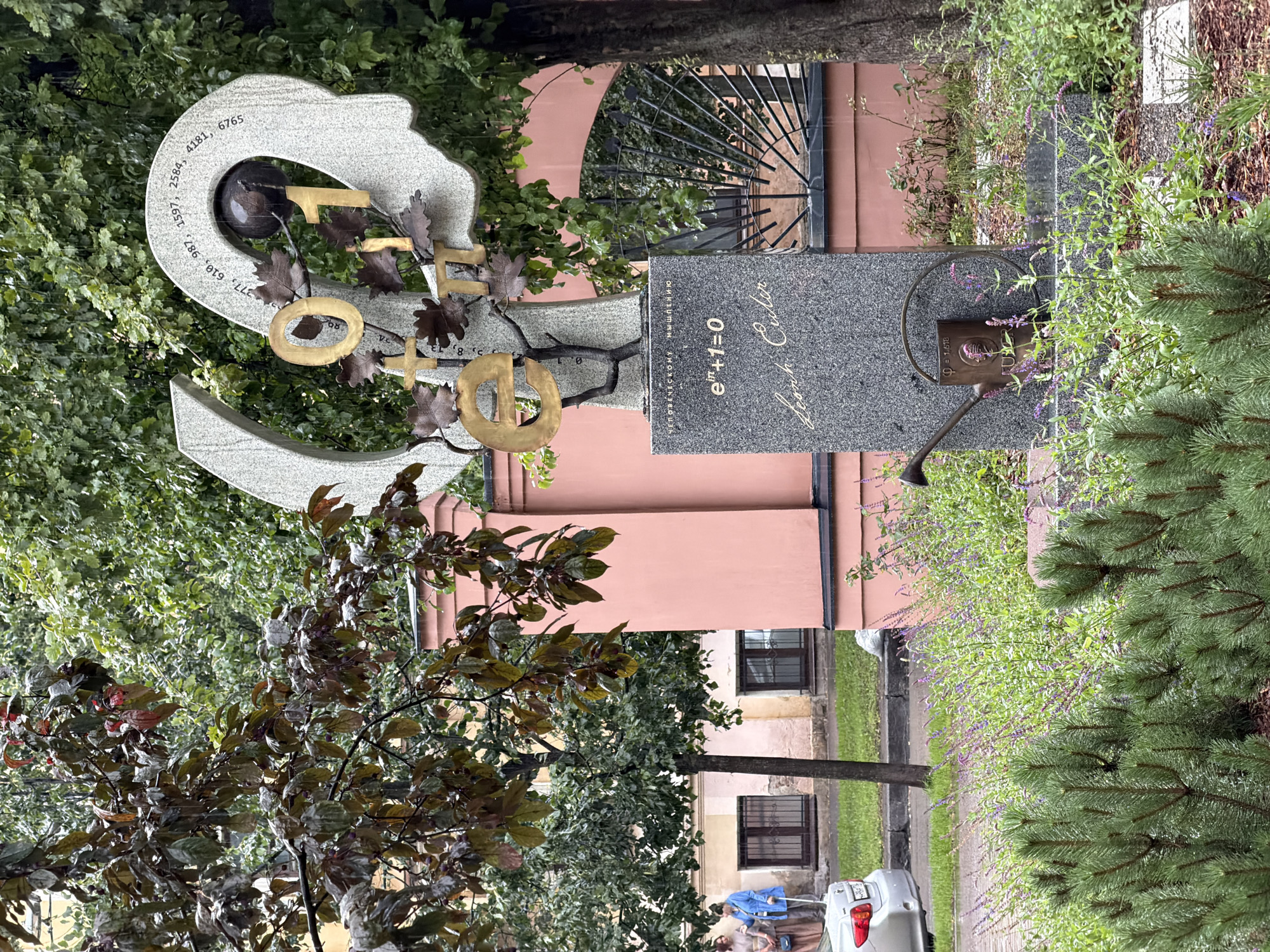 The image depicts a monument dedicated to Leonhard Euler, located in Saint Petersburg, Russia. The monument features a granite base inscribed with the equation e^(iπ) + 1 = 0 and Euler's signature. Above the base, the monument is adorned with golden numbers and mathematical symbols, including "0," "1," and "π," intertwined with stylized oak leaves. The top portion of the monument is shaped like a human head profile, with numbers etched around it, possibly representing dates or significant figures from Euler's life. A dark, spherical object, perhaps a globe, is placed within the head's contour. In front of the monument, a bronze watering can is positioned, with a small plant growing from it, suggesting a dedication to nurturing intellectual growth. The surrounding environment includes lush green trees, a pink building with arched windows, and a street with parked cars. Two women are visible in the background, near the building. The scene is set on a rainy day, with visible raindrops and wet foliage.