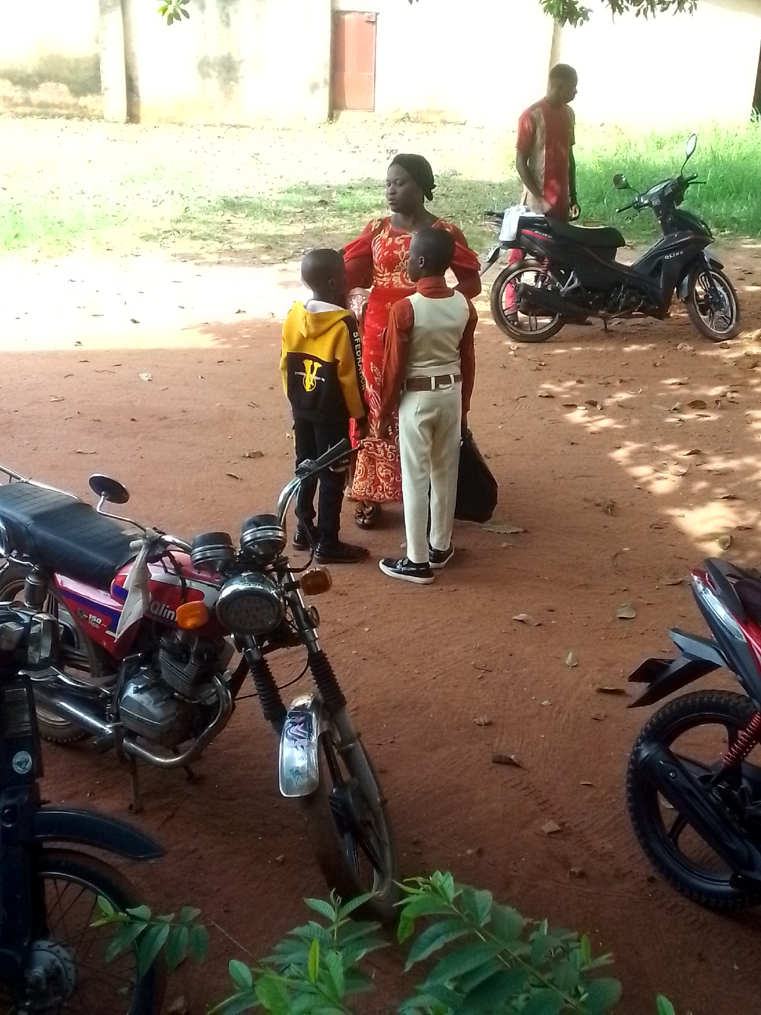 The image depicts an outdoor scene in Unknown, Unknown. A woman in a red patterned dress is standing with two young boys, one in a yellow and black hooded jacket and the other in a white vest and pants. A man in a red shirt is standing near a black motorcycle in the background. There are three motorcycles in the foreground, one red and two black. The ground is reddish-brown dirt. A building with a red door is visible in the background.