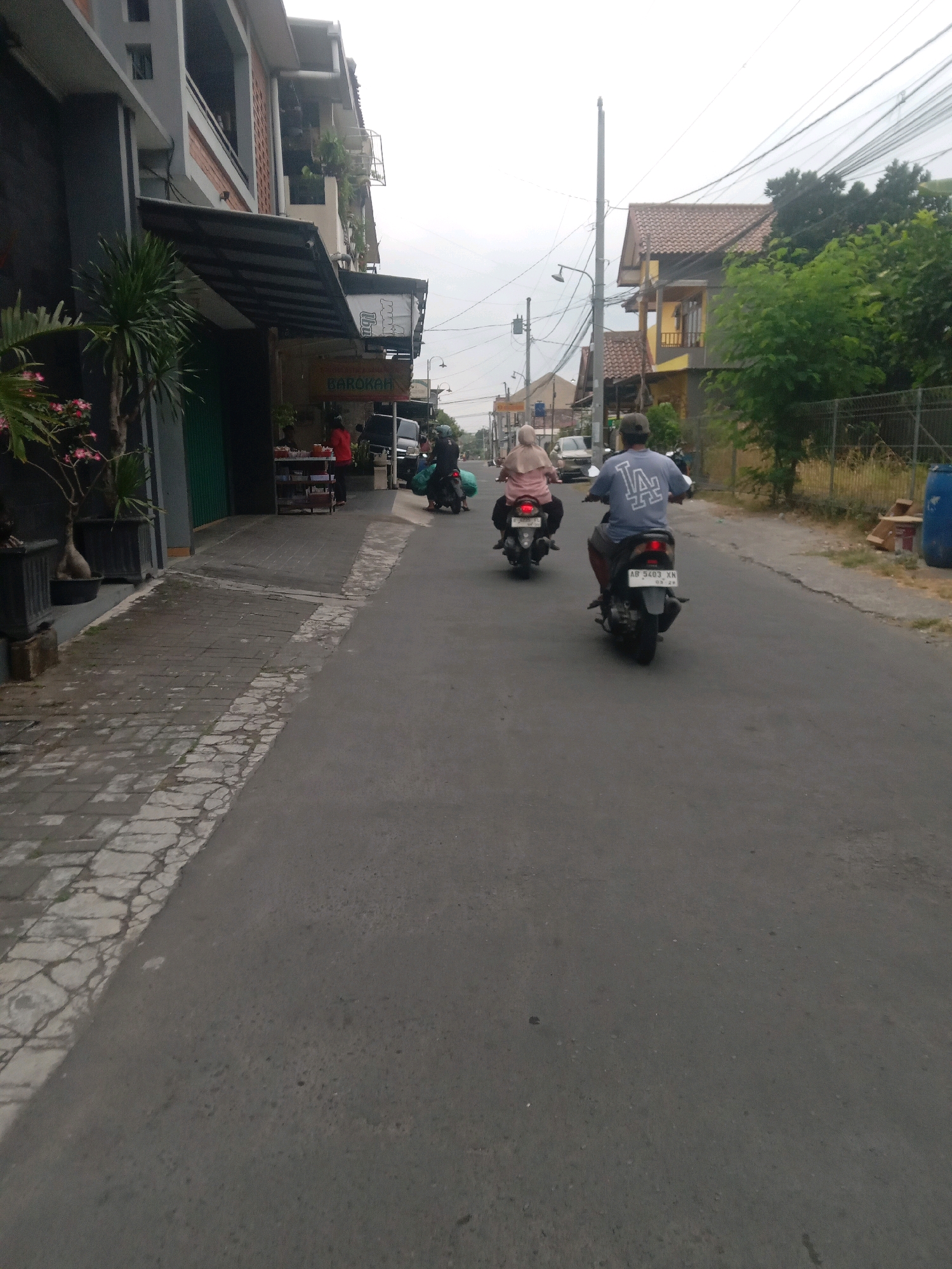 The image shows a street scene in Maguwoharjo, Indonesia. Two people are riding motorcycles on an asphalt road. The person in front is wearing a light pink headscarf and a pink shirt, riding a black motorcycle. The person behind them is wearing a blue t-shirt with a white "LA" logo and a dark cap, riding a black motorcycle with a white license plate reading "AB 5403 XN". To the left of the road, there is a building with a dark facade and a grey upper level, featuring an awning over a sidewalk. Plants in dark planters are visible on the sidewalk. Further down the street, another building with a tiled roof and yellow walls is visible on the right, surrounded by greenery. Overhead power lines crisscross the sky.