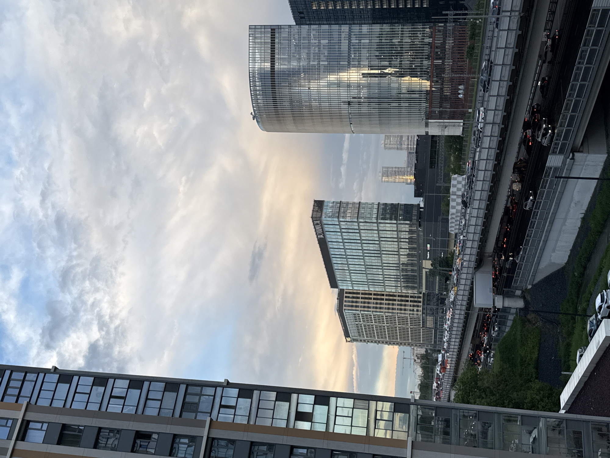 The image depicts a view from a high vantage point in Khimki, Russia. Several modern buildings with glass facades dominate the frame. A multi-lane highway filled with cars runs alongside the buildings. The sky is overcast with various cloud formations.