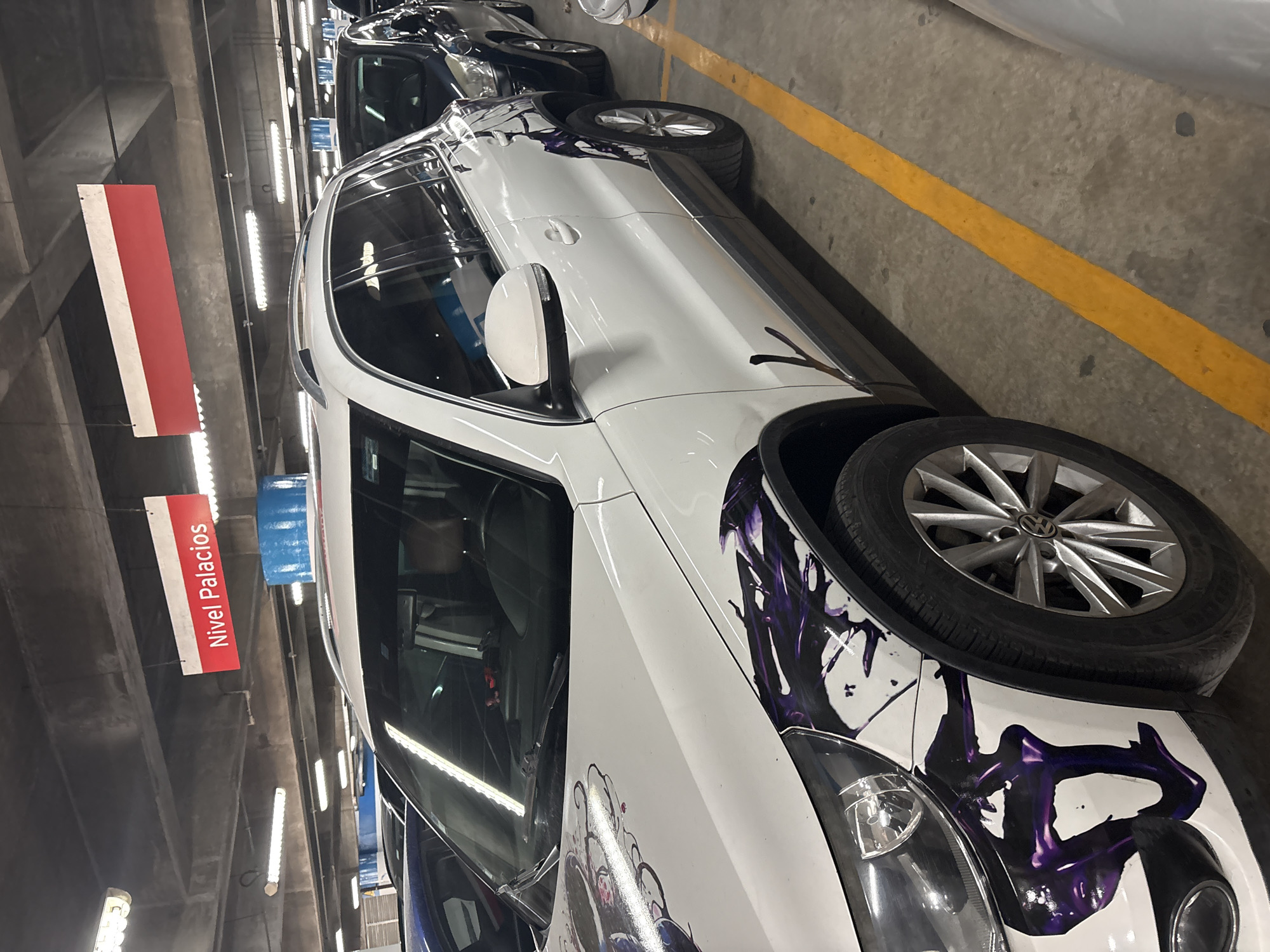 The image shows a white car with purple graffiti-style artwork parked in an underground parking garage. The sign "Nivel Palacios" (Level Palacios) indicates the location is likely within the Palacio de Hierro department store, commonly found in Mexico City. The parking garage is brightly lit with fluorescent lights and has yellow parking lines. Other cars are visible in the background.
