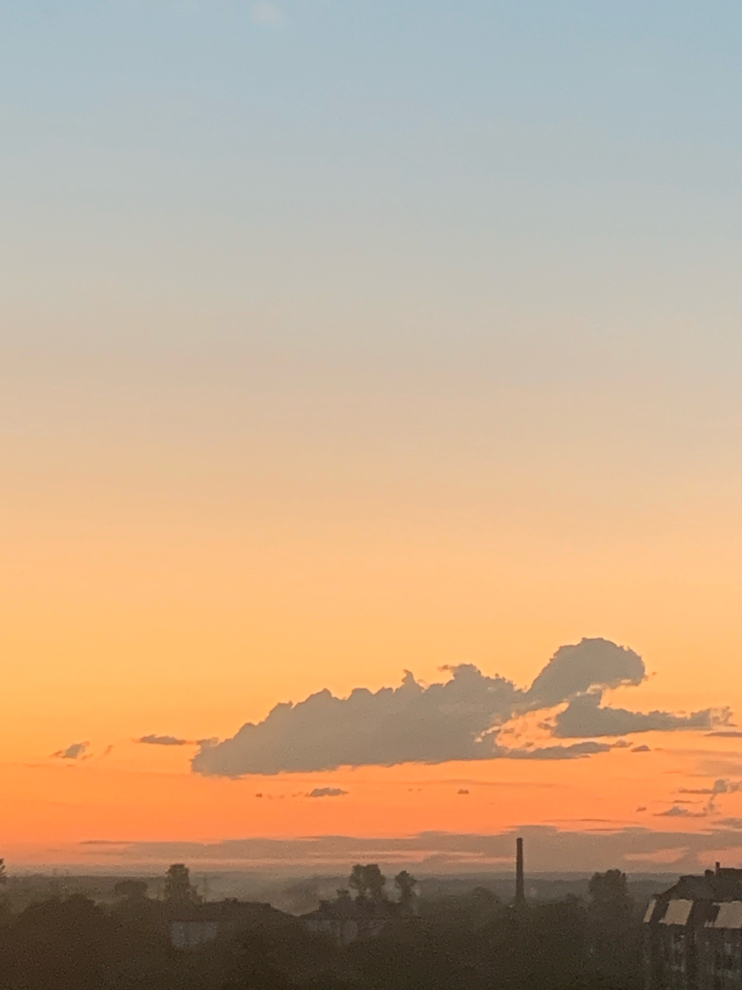 The image depicts a twilight sky over the city of Kolomyia, Ukraine. The sky transitions from a pale blue at the top to a warm orange and pink hue near the horizon, characteristic of sunrise or sunset. A few scattered clouds are visible, with one larger, elongated cloud formation in the mid-sky. The lower portion of the image shows a silhouetted cityscape with rooftops, trees, and a tall, slender smokestack or tower. The overall atmosphere is serene and tranquil, with the fading light casting a soft glow over the urban landscape. There are no people or animals visible in the frame.