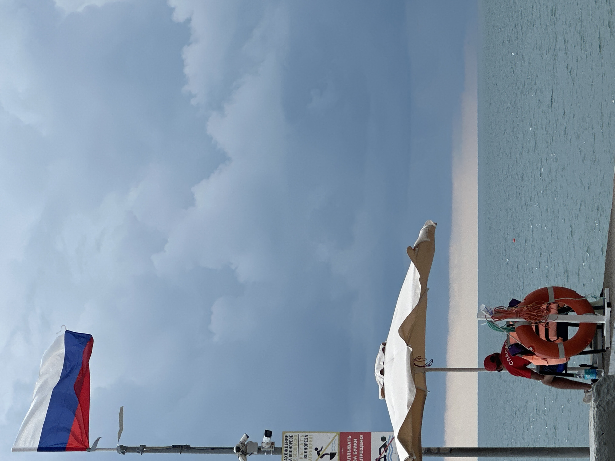 A lifeguard in a red shirt and hat is standing next to a white rack holding two orange life preservers. The lifeguard is looking down at the water. The scene is outdoors with a blue sky and clouds. To the left of the lifeguard, there is a large white umbrella. In the foreground, a Russian flag is waving on a pole. There are also signs and a security camera visible. The water appears to be a calm sea, likely a beach or pier. The time of day is daytime, and the weather is clear with some clouds. The location is Sochi, Russia. Text on a sign reads: "ЗАПРЕЩЕНО" which means "FORBIDDEN" in Russian.