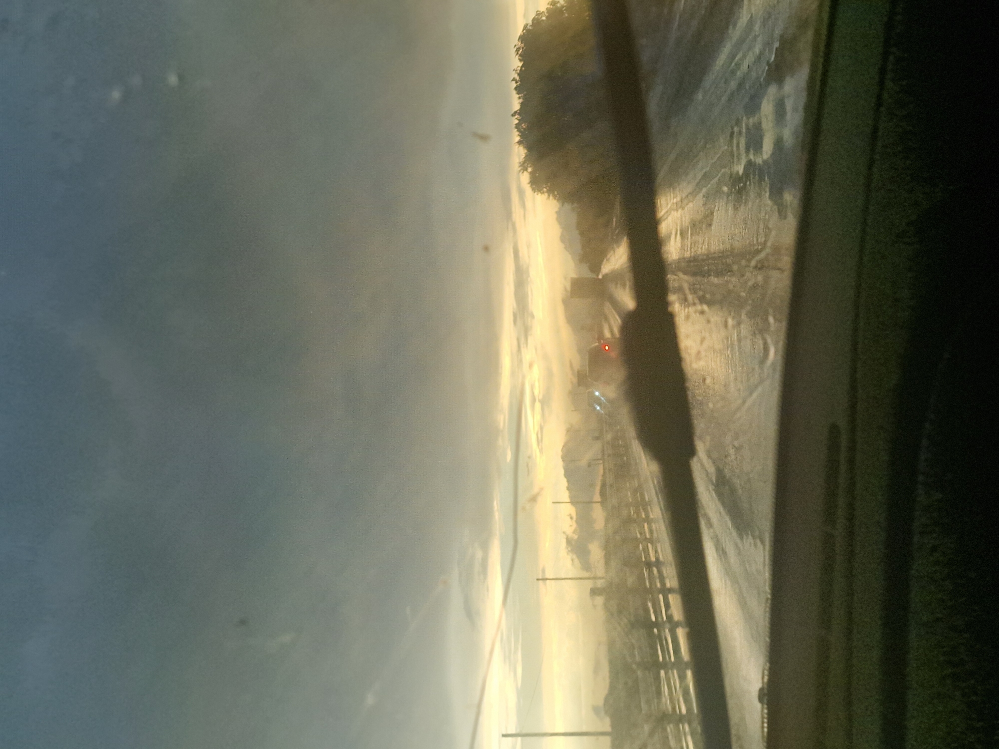 The image captures a view from inside a vehicle, looking outwards through the windshield. The perspective is tilted, giving the impression of motion or an unusual camera angle. The scene appears to be a roadside, likely in Russia, given the location context.
The primary subjects are the road and the sky. The road is wet, reflecting the light and suggesting recent rain or moisture. There are tire tracks visible on the road surface. Along the side of the road, there is a fence-like structure, possibly a guardrail or a field boundary. In the distance, there is a tree line, with some foliage visible. A single red light, possibly from another vehicle, is noticeable on the road ahead.
The sky is overcast with a hazy quality, indicating diffused light. The overall lighting suggests it might be dawn or dusk, as the sky has a soft, luminous glow. The weather appears to be dull or possibly misty, contributing to the muted colors and the wetness of the road.
There is no visible text in the image. The emotion conveyed is one of movement and possibly introspection, given the solitary perspective and the atmospheric conditions. The scene is characteristic of driving through a rural or semi-rural area, potentially on a highway or a main road.