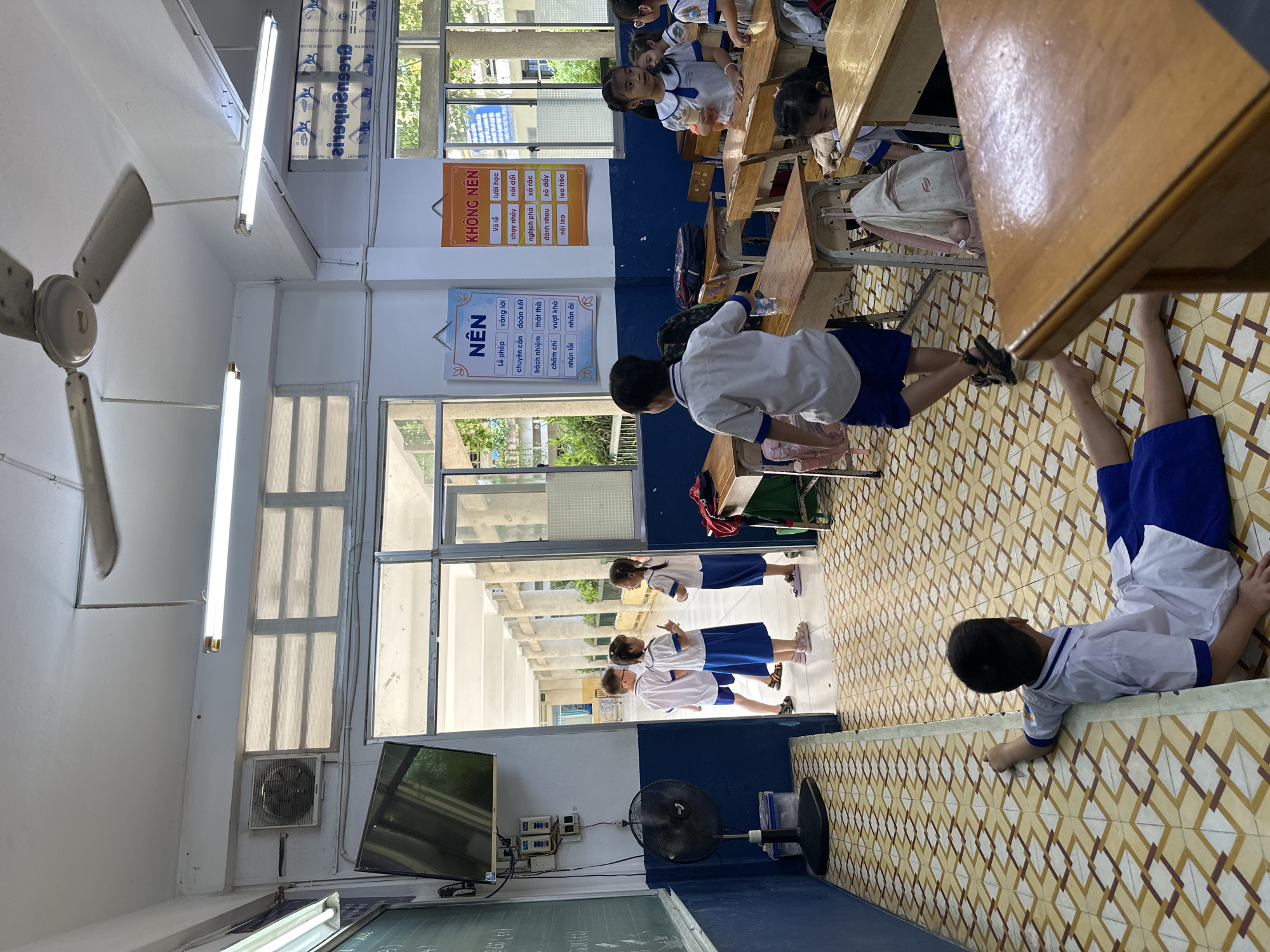The image shows a classroom in Ho Chi Minh City, Vietnam. Several students in white and blue uniforms are present. Some are seated at wooden desks, while others are standing near the window. A sign with Vietnamese text is visible on the wall. The classroom is equipped with ceiling fans, fluorescent lights, and a television on the wall, and the floor is tiled.