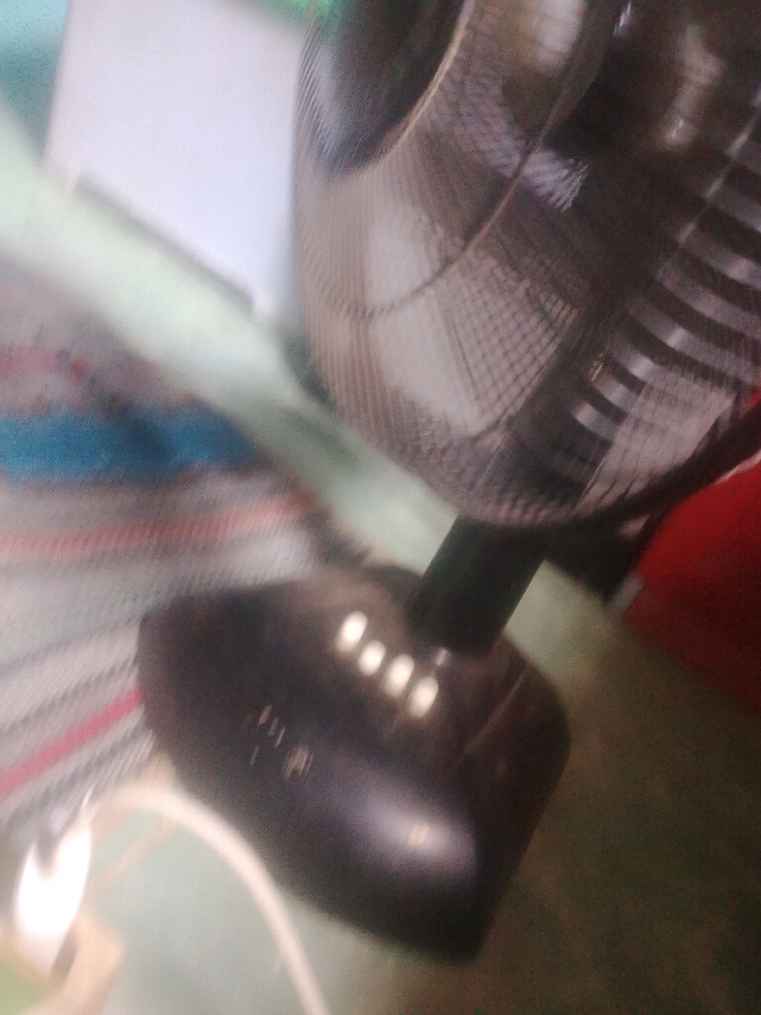 The image shows a close-up, blurred view of a black electric fan with a metal grille. The fan is positioned on a surface that appears to be a striped blanket with red, white, and blue stripes. A white cord is visible extending from the base of the fan. In the background, a white surface, possibly a wall or a piece of furniture, is partially visible. The overall scene is dimly lit and appears to be indoors. No people are present in the image. The location in Kecamatan Haurgeulis, Indonesia is not discernible from the visual information provided in the image.