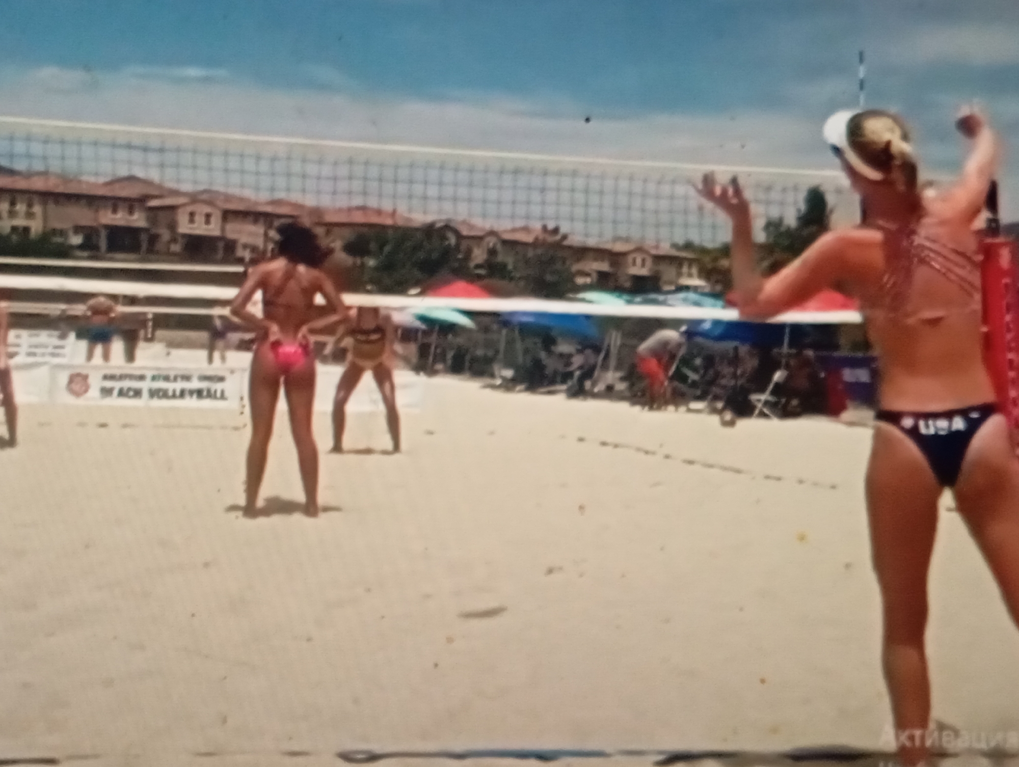 A beach volleyball game is in progress on a sandy court. A woman in a red bikini is positioned at the net, facing away from the camera. Her hands are raised, and she appears to be in the process of serving or blocking. To her left, another woman in a pink bikini stands with her hands on her hips, looking towards the net. Further back, on the other side of the net, a woman in a yellow bikini is also visible, as is a man in orange shorts. In the background, buildings with Spanish-style architecture are visible, along with trees and a blue sky. A banner with text that includes "BEACH VOLLEYBALL" is displayed near the net. The scene is brightly lit, suggesting it is daytime. The location of Sofiivska Borshchahivka, Ukraine is not discernible from the image.