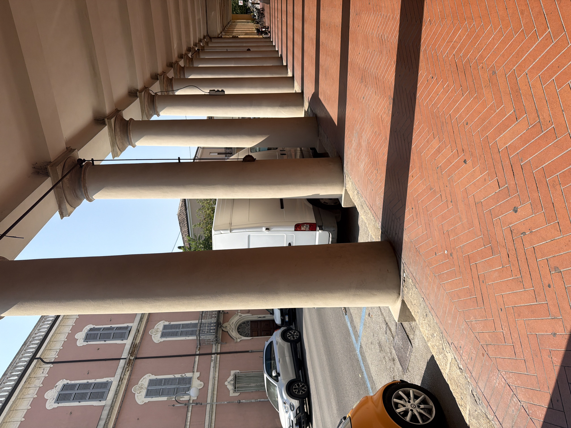 The image depicts a street scene in Santarcangelo di Romagna, Italy, characterized by a long colonnade with prominent rounded columns. The colonnade casts distinct shadows on the red brick pavement, laid in a herringbone pattern. In the foreground, the wheel and lower portion of a small orange car are visible on the left. Behind it, a silver SUV is parked on the street, alongside a white van. The buildings lining the street are visible beyond the colonnade, featuring a pink facade with shuttered windows and decorative architectural elements. The sky is clear and blue, indicating a sunny day. In the distance, at the end of the colonnade, several parked motorcycles and bicycles suggest a public area.