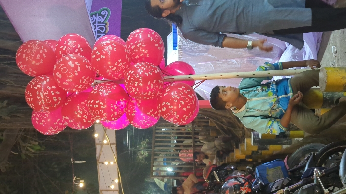 The image depicts an outdoor evening or night scene, centered around a display of numerous red balloons. The balloons are primarily red with white "LOVE" text and small heart patterns printed on them. They appear to be tied together and held aloft by a wooden pole or staff.
A young boy is visible on the right side of the frame, wearing a light blue and white zippered jacket and brown pants. He is sitting on a yellow and black striped barrier or bollard, with another similar barrier behind him. He holds the wooden pole attached to the balloons and is looking upwards. His expression seems calm and attentive.
Behind the boy, a man with a beard and long, dark hair is standing. He is wearing a grey, long-sleeved shirt. He appears to be looking towards the left side of the frame, possibly at the balloons or interacting with someone out of view.
The background is dimly lit, suggesting it is either evening or nighttime. String lights or small bulbs are visible in the lower left and center background, adding to a festive ambiance. In the upper left background, there's a purple banner or sign with text written in Bengali script. Below the boy and the balloons, several parked motorcycles or scooters are partially visible. The ground appears to be asphalt or concrete. The overall scene suggests a street, market, or event gathering.