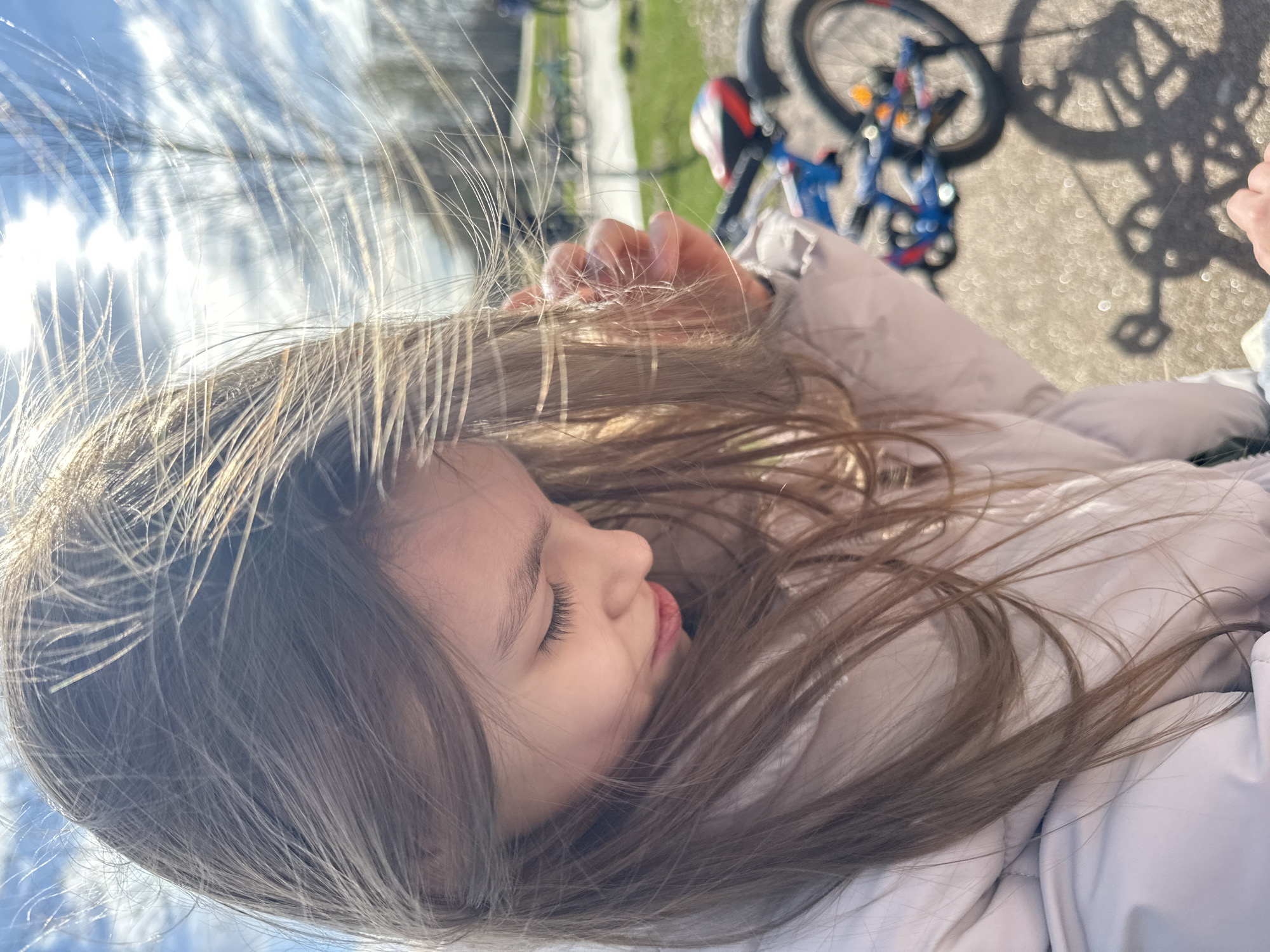 A close-up, tilted perspective shows a young girl with long brown hair lying down. Her eyes are closed, and she appears to be resting. A hand is visible near her head. In the blurred background, a bicycle, a paved path, and some greenery are discernible. The location is likely in a park area in Fryazino, Russia.