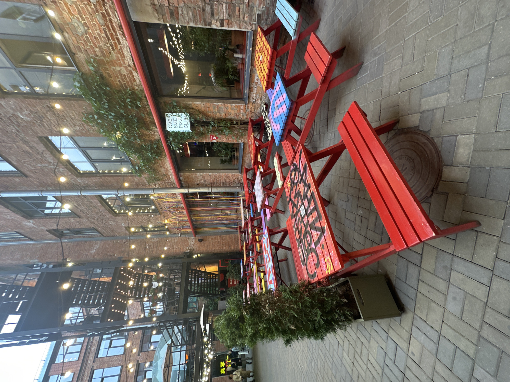 The image shows an outdoor seating area in what appears to be a courtyard or alleyway. The area is lined with brick buildings with various windows and architectural details. String lights are visible above. The seating consists of small, brightly painted red tables and benches. The tables are decorated with different patterns and designs. A row of potted evergreen plants separates the tables from the pedestrian walkway.
The walkway is paved with gray bricks. A manhole cover is visible in the foreground. In the background, there are people walking. There is a sign for "OVER SIZE PIZZA CLUB" on one of the buildings.
Given the architectural style and the signage in Cyrillic script, it is highly probable that the location is in the city of Saint Petersburg, Russia.