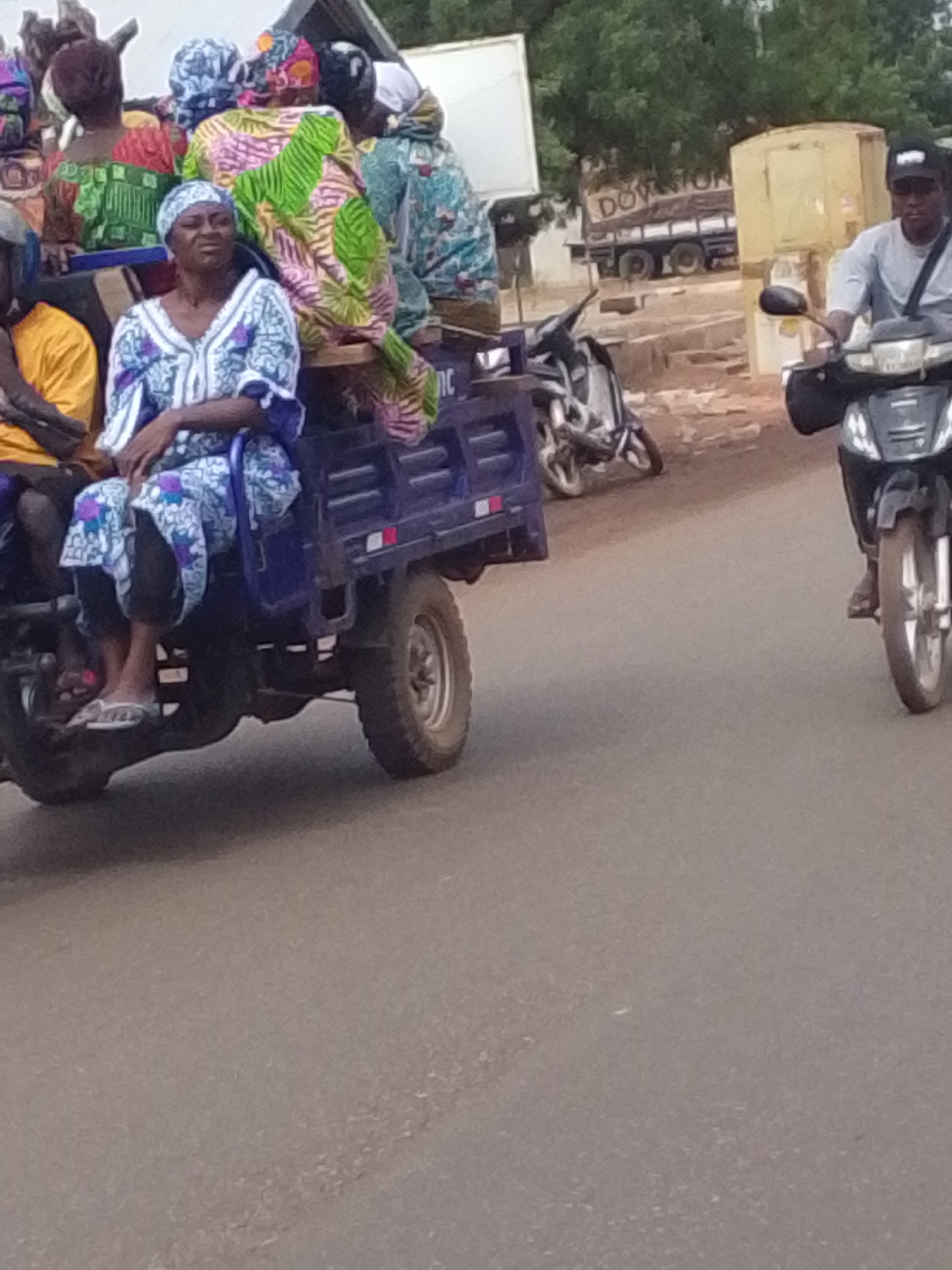 The image displays a street scene in Tamale, Ghana, featuring two vehicles on a paved asphalt road. In the left midground, a blue three-wheeled cargo tricycle, equipped with a metal bed, is transporting at least seven individuals. A woman prominently occupies the front-left passenger area of the tricycle, dressed in a white and blue patterned tunic and headscarf, wearing white flip-flops. Behind her, other women are seated in the cargo bed, adorned in brightly colored traditional patterned clothing, including green, pink, and blue designs. A person in a yellow shirt is partially visible on the far left.
In the right midground, a man operates a black scooter-style motorcycle, moving parallel to the tricycle. He wears a light-colored short-sleeved shirt, a dark baseball cap, and a dark bag slung across his chest.
The background consists of green foliage from trees and various structures, including a white rectangular sign. A parked truck, partially obscured, shows the word "DOWNTOWN" on its side. Another motorcycle is parked on the roadside between the two moving vehicles. The environment is consistent with an urban or semi-urban setting.