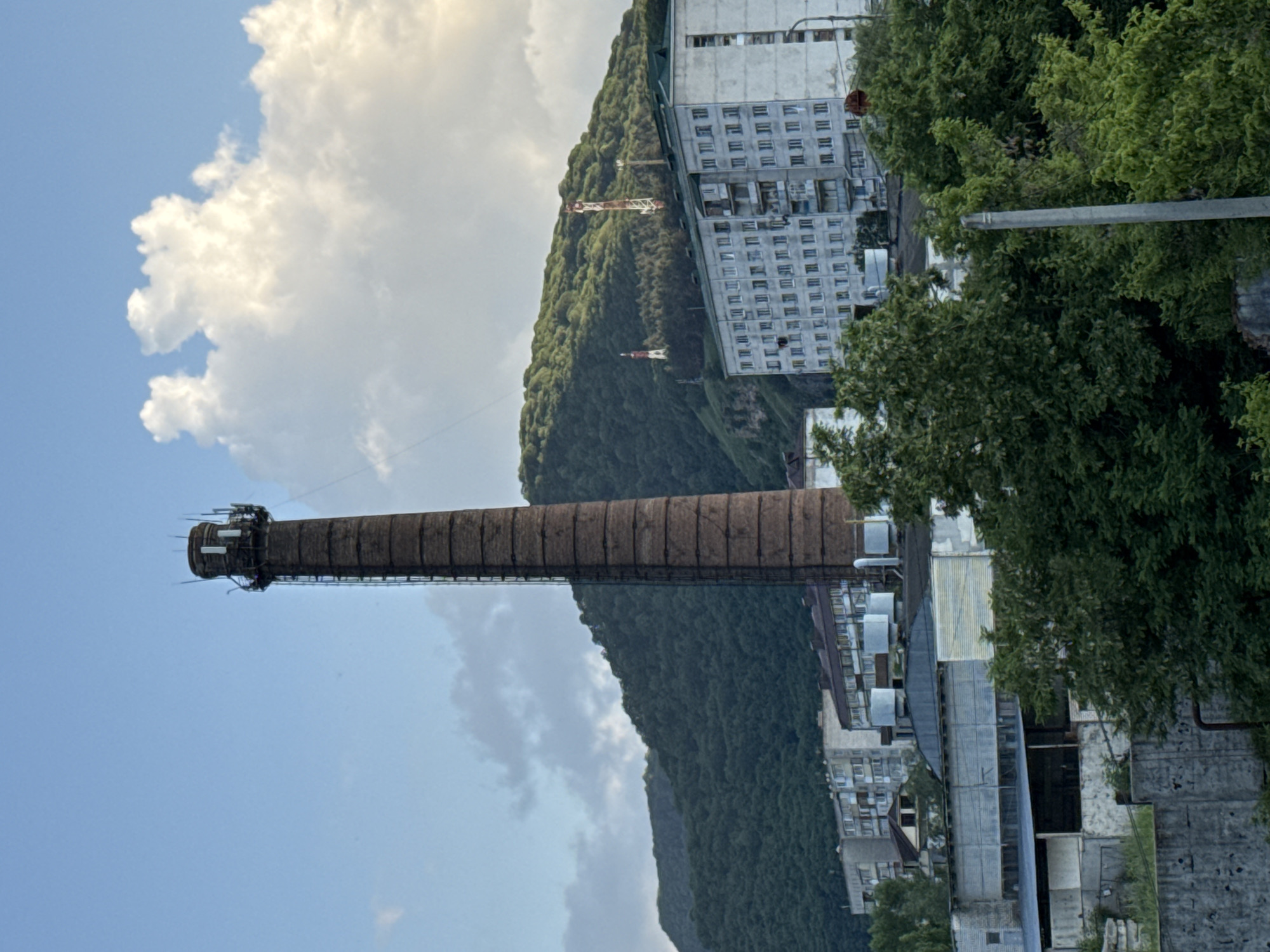 The image depicts an industrial scene in the city of Karachayevsk, Russia, featuring a tall, brick smokestack as the central element. The smokestack, showing signs of age and wear, is positioned against a backdrop of a lush, green, tree-covered hillside. In the mid-ground, several multi-story residential buildings with balconies are visible, along with some industrial structures. Two communication towers with red and white markings are situated on the hillside, indicating the presence of telecommunication infrastructure. The sky is partly cloudy, with large, white cumulus clouds contrasting with patches of blue sky. The overall impression is one of an industrial area nestled within a natural, mountainous landscape.