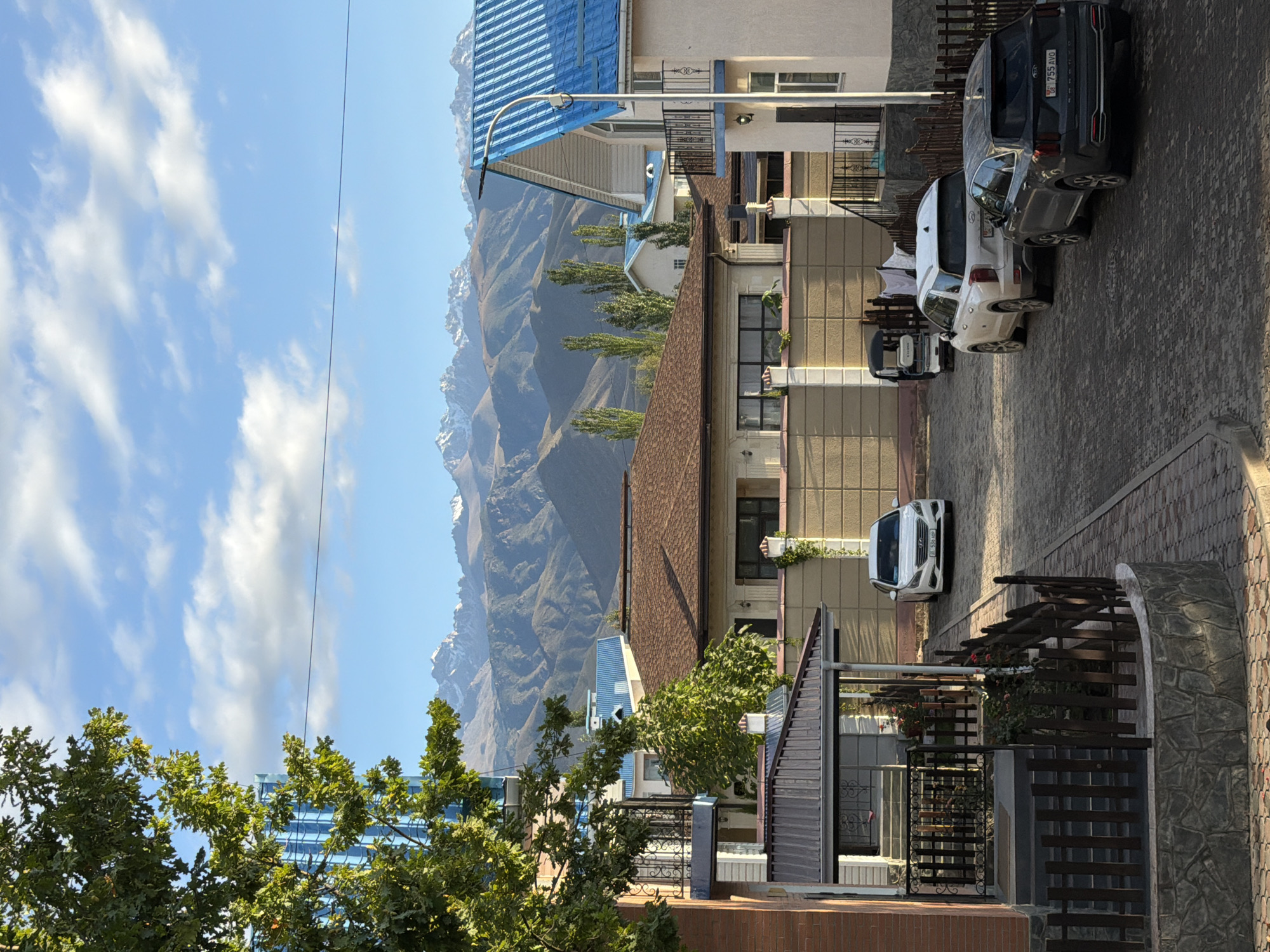 The image depicts a street scene in Cholpon-Ata, Kyrgyzstan, with several houses and vehicles. The foreground features a paved street with dark gray cobblestones. To the left, a brick building with a blue vertical grate is partially visible, with lush green foliage in front of it. Further down the street on the left, a house with a dark metal roofed porch is present, with a small dark wooden fence and stone base.
In the mid-ground, a light-colored sedan is parked on the right side of the street. Beyond it, a white SUV and a smaller white vehicle resembling a golf cart are parked in front of a beige textured wall. Several tall, slender trees are planted behind this wall.
The background is dominated by a residential area with houses featuring brown and blue roofs. In the far background, a majestic mountain range with snow-capped peaks is visible under a bright blue sky with scattered white clouds. The overall atmosphere is one of a quiet, sunlit day in a suburban or semi-rural setting, with a clear view of natural landscapes.