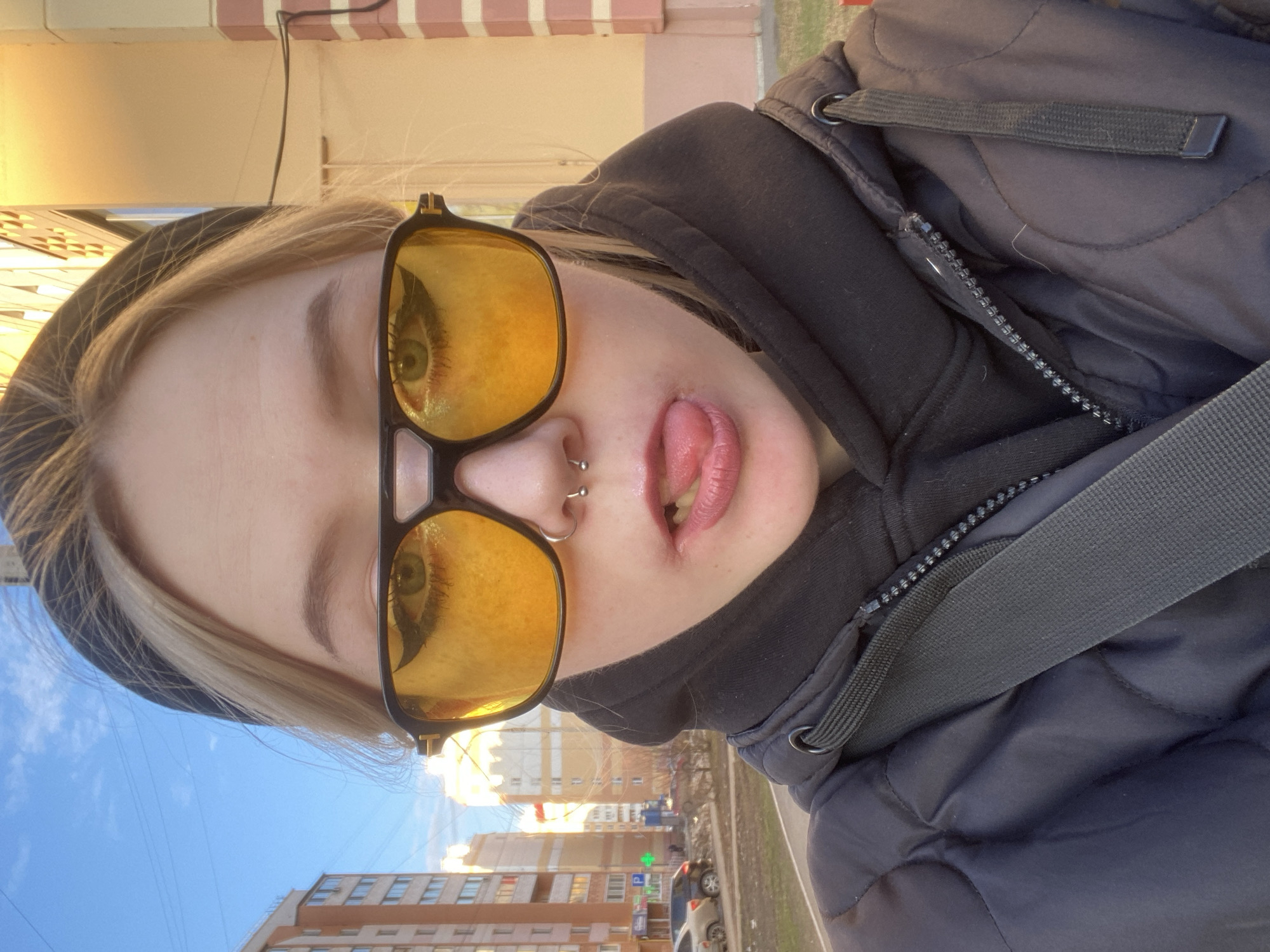 A young individual with blond hair, wearing yellow-tinted sunglasses, a dark-colored hat, and a dark puffy jacket is pictured in a selfie. The individual has facial piercings in the nose and is sticking out their tongue. The background shows a cityscape, with several residential buildings. A pharmacy sign is visible. Based on the architecture and the pharmacy signage, the location is likely Izhevsk, Russia.