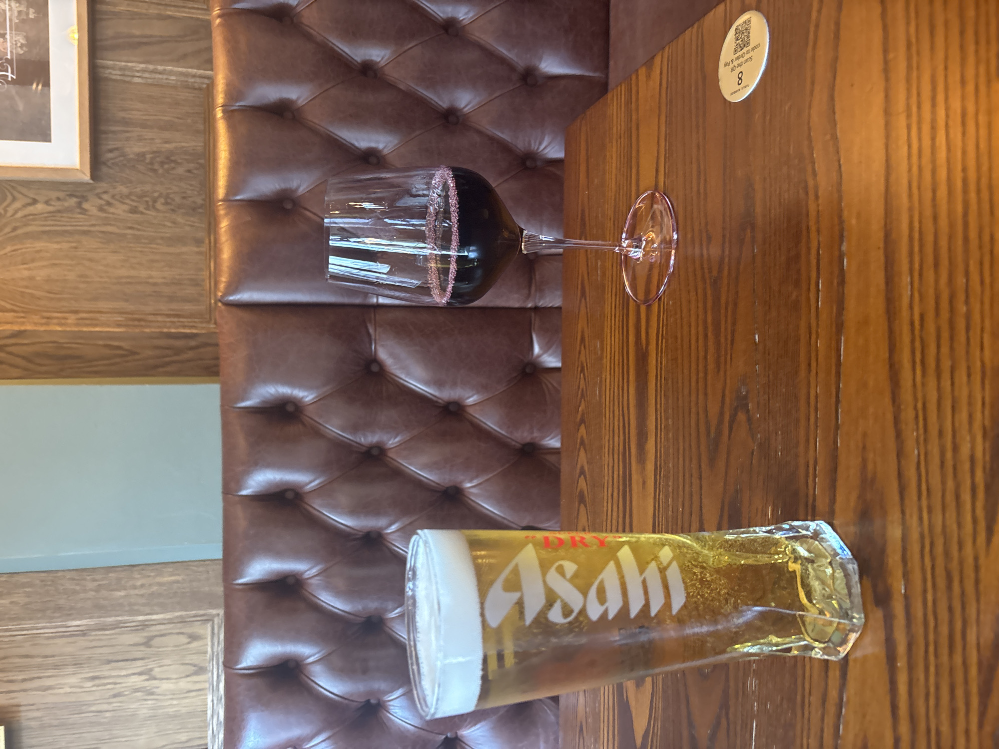A slightly tilted overhead shot captures a table in an indoor setting, with two beverages placed on it: a tall, cylindrical glass of Asahi beer with a thick white foam head and a wine glass with a dark-colored liquid and a decorative, pink rim. The table is made of light brown wood, showing vertical grain. To the left, a section of a brown, tufted, leather-like bench is visible, along with a glimpse of a light blue wall and light brown wooden paneling. A small, circular sticker with a QR code and the number "8" is adhered to the table. Based on the presence of Asahi beer and the general aesthetic, the location could be a restaurant or bar in London, United Kingdom, although there are no definitive landmarks to confirm this.