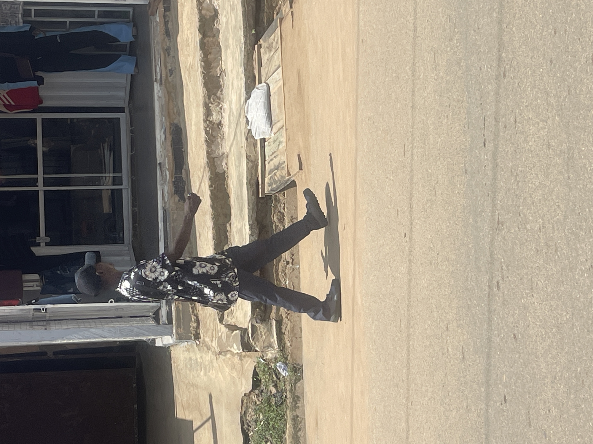 A young boy wearing a black baseball cap and a floral print shirt is walking across a dusty road in Abuja, Nigeria. He has a dark bag slung over his shoulder. The boy is walking from left to right in the frame, with his body angled towards the viewer. In the background, there is a building with a storefront that has a metal security gate. Steps lead up to the storefront, and a white bundle of what appears to be clothing or fabric is placed on a wooden plank structure next to the steps. The ground is a mix of concrete and dirt, with some patchy green vegetation visible near the building. The scene appears to be taking place during daylight hours, with shadows cast by the boy and the structures.