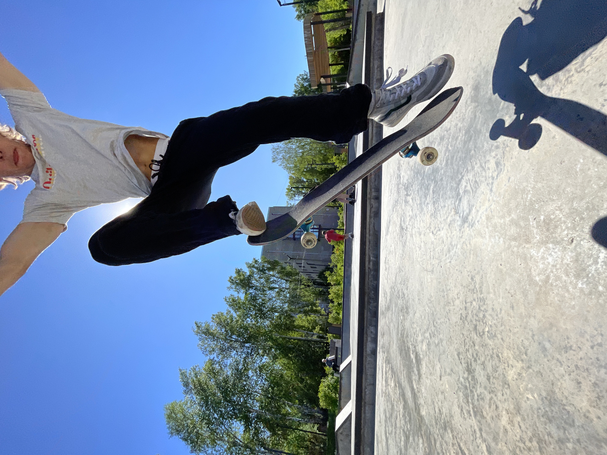 A person is captured mid-air, performing a skateboard trick in a public outdoor skatepark. The skateboarder, dressed in a gray t-shirt and black pants, is positioned above the concrete ground, with their skateboard beneath their feet. The sun is bright, creating a lens flare effect and casting a shadow of the skateboarder and their board onto the ground. In the background, trees and buildings are visible, suggesting an urban or suburban setting. The skatepark features concrete ramps and structures. The image is taken from a low angle, emphasizing the height of the jump.