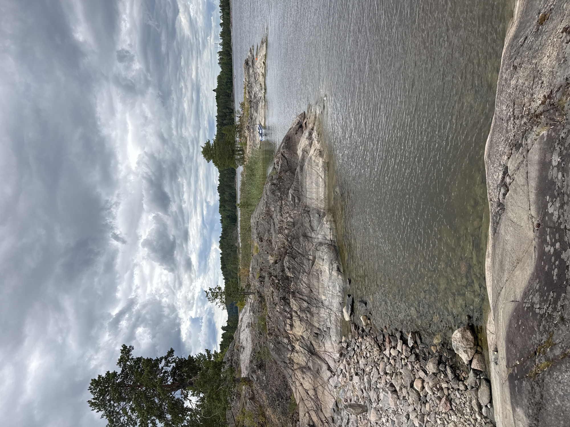 A rocky shoreline in Sorola, Russia, is depicted under a heavily clouded sky. The foreground features large, weathered rocks with a scattering of smaller stones leading down to the water's edge. The water itself is calm with visible ripples. In the midground, a small, white and blue boat is moored near a rocky islet dotted with sparse trees. Beyond the islet, a dense forest lines the horizon. The overall atmosphere is somber, likely due to the overcast conditions. There are no people or animals visible in the image.