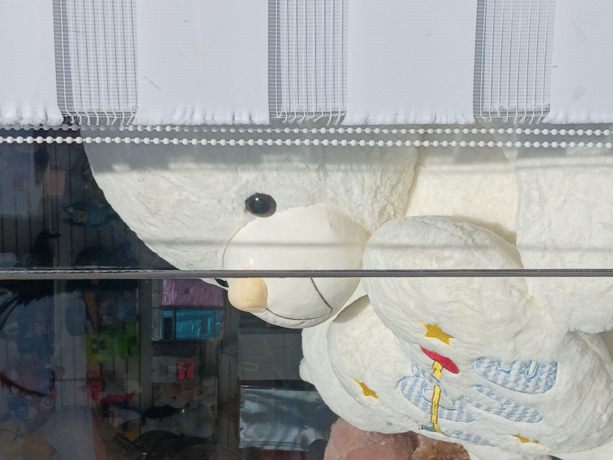 The image captures a large, fluffy white teddy bear viewed through a glass pane, possibly a shop window or display. The bear is oriented on its side, occupying a significant portion of the frame. It has a dark, round eye and a light brown snout. On its white, soft belly, there are embroidered designs including several yellow stars, blue cloud-like shapes with a textured pattern, and a central figure with red and yellow elements, resembling a stylized candle or a character with a hat and staff.
Above the teddy bear, white vertical blinds with a fine mesh pattern are visible, featuring a decorative white beaded chain along their lower edge, suggesting they are partially drawn. A dark, horizontal bar or frame runs across the mid-section of the image, separating the blinds and the bear from the background details.
Through the glass and behind the bear, a blurred background reveals shelves or display units holding various colorful objects. Rectangular boxes in shades of pink, blue, and green are discernible, hinting at a retail environment, such as a toy store or gift shop. There is no discernible activity taking place, as it is a static shot of objects. No people or visible text are present in the image. The lighting appears bright, possibly from natural daylight or strong indoor illumination. While the image shows no specific time of day or weather cues, the scene depicts an indoor setting, likely in Ciudad Guayana, Venezuela, as provided by the context.