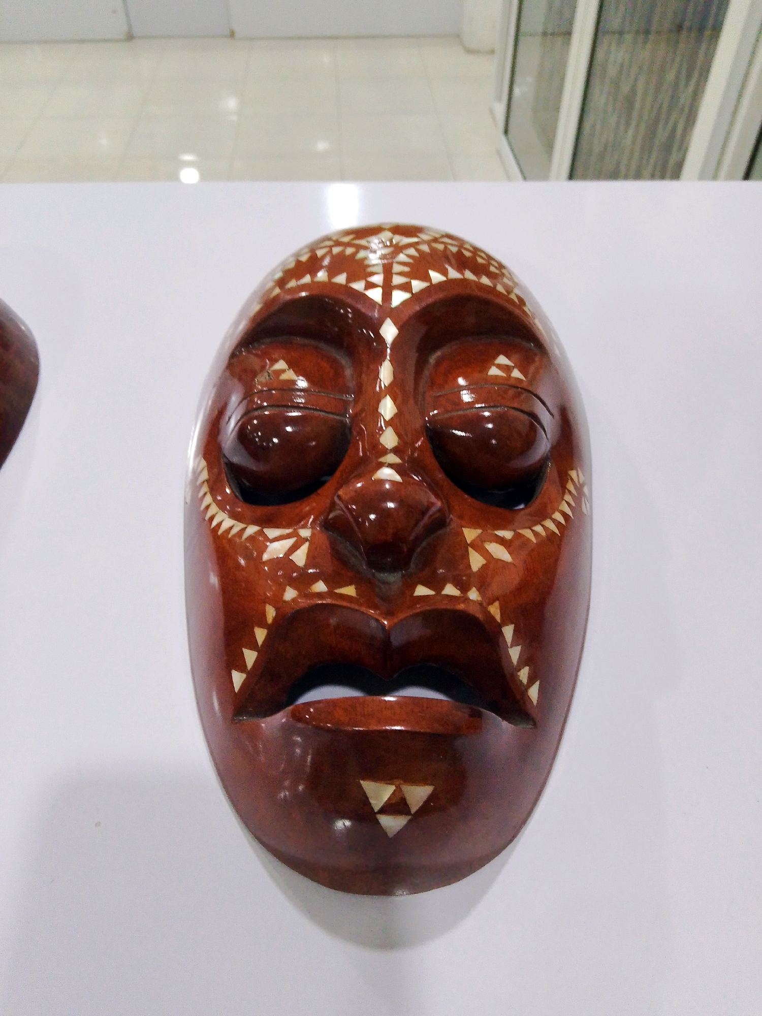A close-up, front-facing shot shows a traditional wooden mask with a reddish-brown, polished finish. The mask is adorned with intricate white triangular and diamond-shaped inlays, forming decorative patterns around the eyes, nose, and mouth. The eyes are depicted as dark, hollowed-out openings, and the lips are slightly parted, revealing a dark interior. The mask rests on a flat, white surface. The background is blurred, showing a tiled ceiling and glass windows, suggesting an indoor display or exhibition space. The mask is centrally positioned, occupying the majority of the frame. The image does not provide any information about the city of Bunkate, Indonesia.