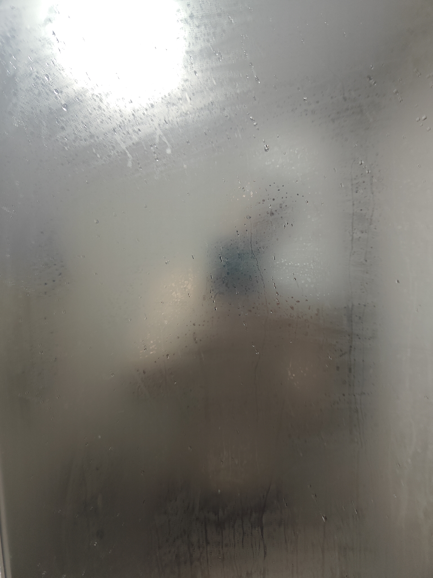 The image shows a foggy window with water droplets. A bright light source is visible in the upper left corner, suggesting it might be daylight or a strong artificial light. The lower part of the image is darker and appears to have streaks of water. The overall scene is abstract and lacks specific details that would allow for identification of objects or people. The location in the city of Shkurupiivka, Ukraine, cannot be determined from the visual information provided in the image.