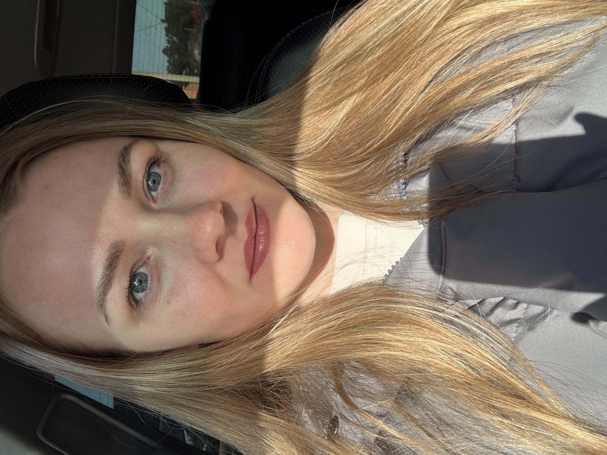 A selfie of a fair-skinned woman with light blonde hair is angled to the left, presumably taken in a car. The woman has light-colored eyes and is wearing lip gloss. A shadow crosses her face from the upper left. She is wearing a gray jacket over a light-colored top. The background shows a dark car interior with a glimpse of a building and trees visible through the window. There is no indication of the city or location.