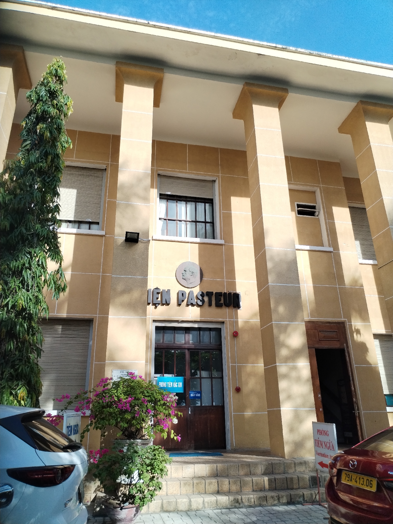 The image depicts the exterior of a building in Nha Trang Ward, Vietnam, specifically the "IEN PASTEUR" (Pasteur Institute). The building has a beige facade with architectural features including columns and a flat roof. There are windows with closed blinds on the first and second floors. A sign above the main entrance door reads "PHÒNG TIÊM VẮC XIN" (Vaccination Room). To the left of the entrance, there's a tree and potted pink bougainvillea plants. Parked cars flank the building. A sign pointing towards the vaccination area is visible next to a parked vehicle, with the license plate 79A-413.06, on the right. There is also a small portrait on the building. The sky is blue.
