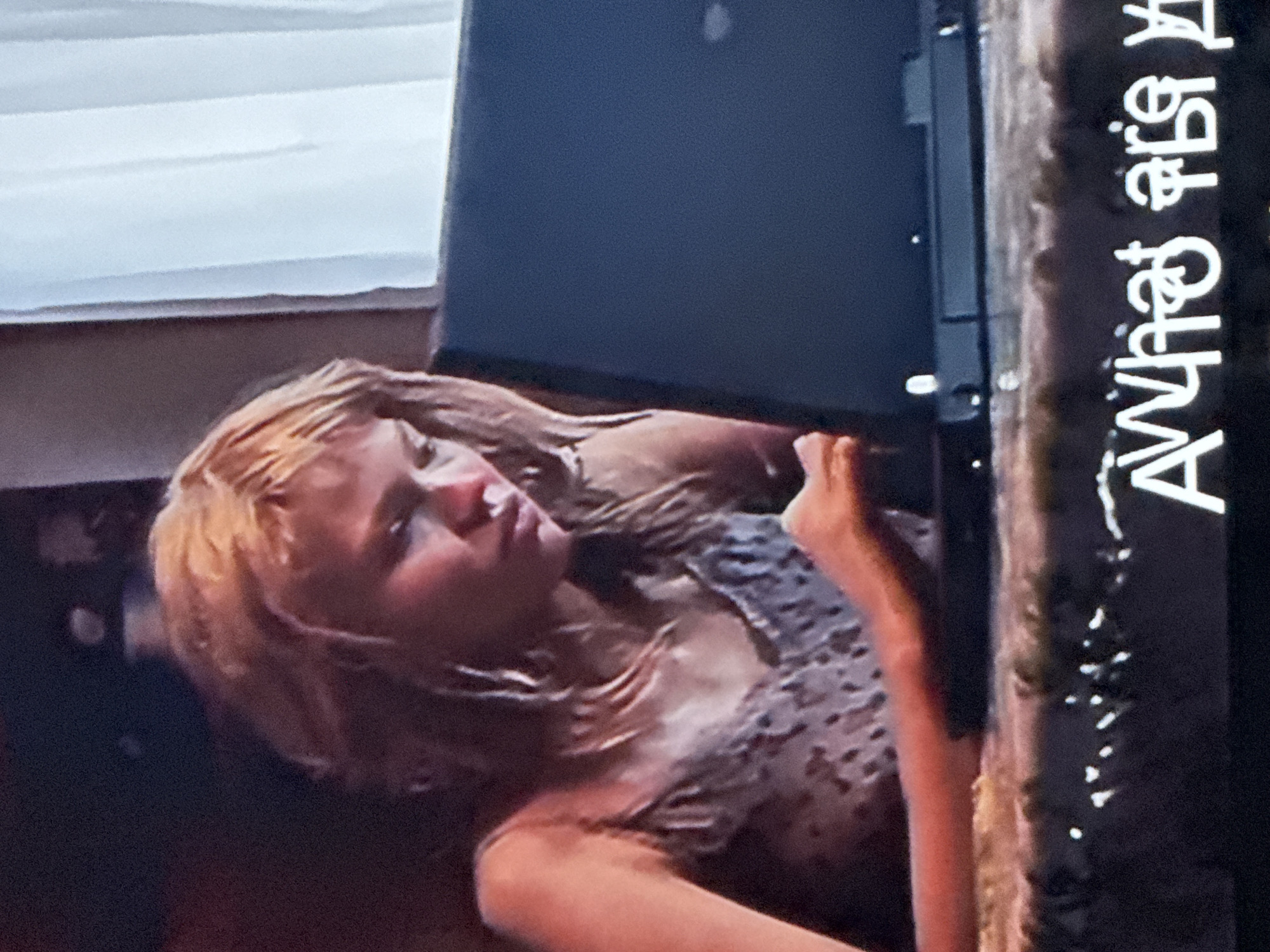 A woman with light-colored hair is lying down. Part of a white blind is visible in the background. A dark object resembling a laptop is positioned above her. A vertical surface with the text "What are you" is present on the right side of the image. The location is not discernible from the visual information.