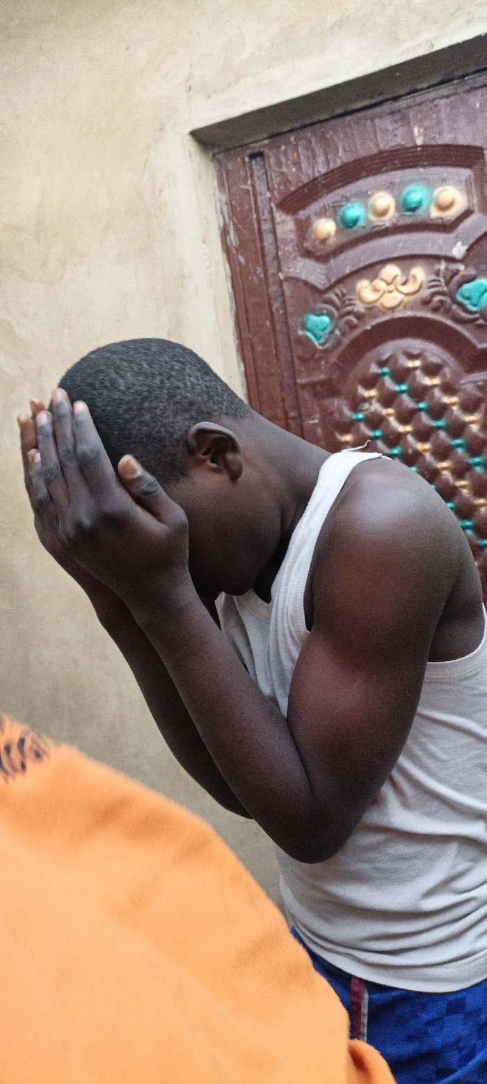 The scene in Kaura Goje, Nigeria, depicts a young male with dark skin and short, dark hair, positioned with his head bowed forward and hands covering his face. He is wearing a white sleeveless undershirt and blue shorts featuring a faint checkered pattern. His hands are clasped together, palms pressed against his forehead and eyes. Behind him, to the left, is a light beige or tan wall with a smooth, possibly stuccoed texture. To his right, a dark reddish-brown wooden door or doorframe is visible, featuring ornate, embossed or carved decorative patterns in teal and orange/gold colors. A portion of an orange textile object or garment is present in the lower-left foreground, partially obscuring the view, suggesting another presence or item nearby.