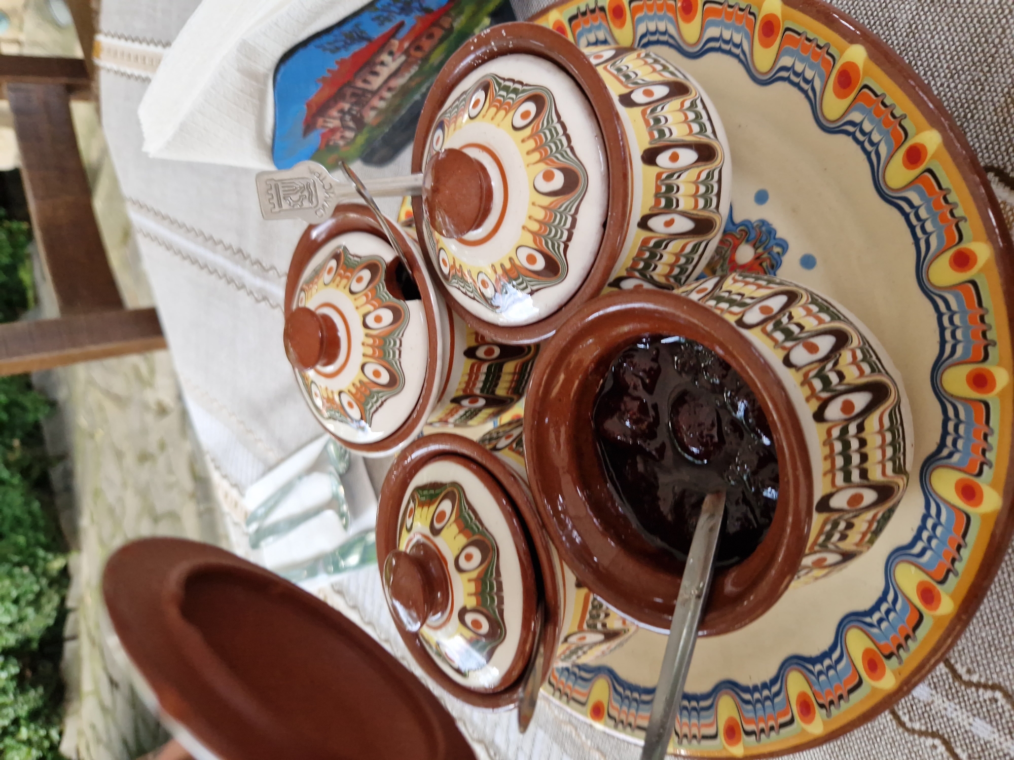 The image depicts a close-up view of a table setting, likely in Panagyurishte, Bulgaria, featuring traditional Bulgarian pottery. The focus is on a decorative ceramic platter holding several small, lidded ceramic bowls. One bowl in the foreground is filled with a dark, jam-like substance and has a spoon resting inside it. Two other lidded bowls are visible, each adorned with intricate, colorful geometric patterns in shades of brown, yellow, green, and white, featuring eye-like motifs. The lids also share this distinctive decorative style. To the right of the bowls, a stack of white napkins is present, with a decorative element that appears to be a small souvenir depicting a building, possibly a historical structure from Panagyurishte. A silver spoon with an embossed emblem, resembling a crest or coat of arms, is placed in one of the lidded bowls. A brown ceramic lid is held in the upper left corner of the frame, partially obscuring the background. The table is covered with a light-colored, textured cloth, and in the blurred background, hints of greenery and wooden furniture suggest an outdoor or semi-outdoor setting, possibly a restaurant or a veranda. The overall impression is one of a traditional culinary presentation, likely offering preserves or condiments, with an emphasis on the ornate craftsmanship of the pottery.