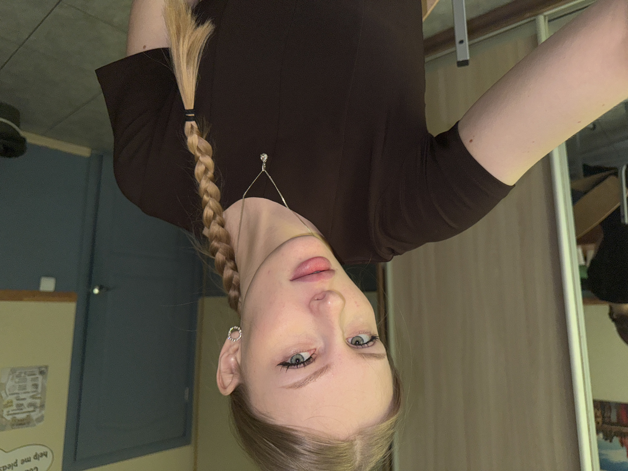 A young, fair-skinned woman with braided blonde hair, wearing a dark brown, long-sleeved top and a delicate silver necklace, is visible from a slightly angled, upside-down perspective. She has light blue eyes and appears to be wearing eyeliner. Her right ear is adorned with a silver hoop earring. The background shows a room with light-colored walls, a blue door, and a white-framed mirror reflecting part of the room. A framed paper with a map and text "help me please" is posted on the wall. Based on the text the location is the city of Perm, Russia.