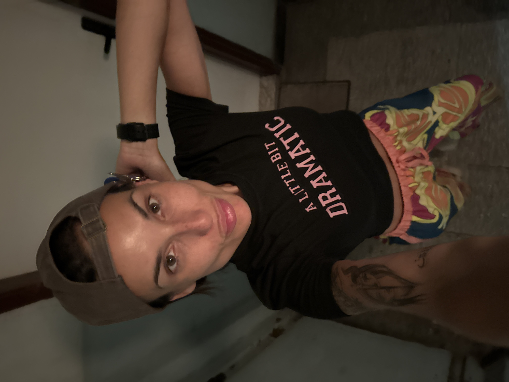 A woman wearing a black t-shirt with pink lettering that says "A LITTLE BIT DRAMATIC" and colorful patterned shorts, along with a grey baseball cap, is standing in an indoor setting. She is looking towards the camera with her mouth slightly puckered. Her left arm is raised, and she is touching the back of her head. A watch is visible on her right wrist. A tattoo is present on her left forearm. The environment appears to be a room with tiled flooring and doorways visible in the background, suggesting an interior space within the city of Pleven, Bulgaria.