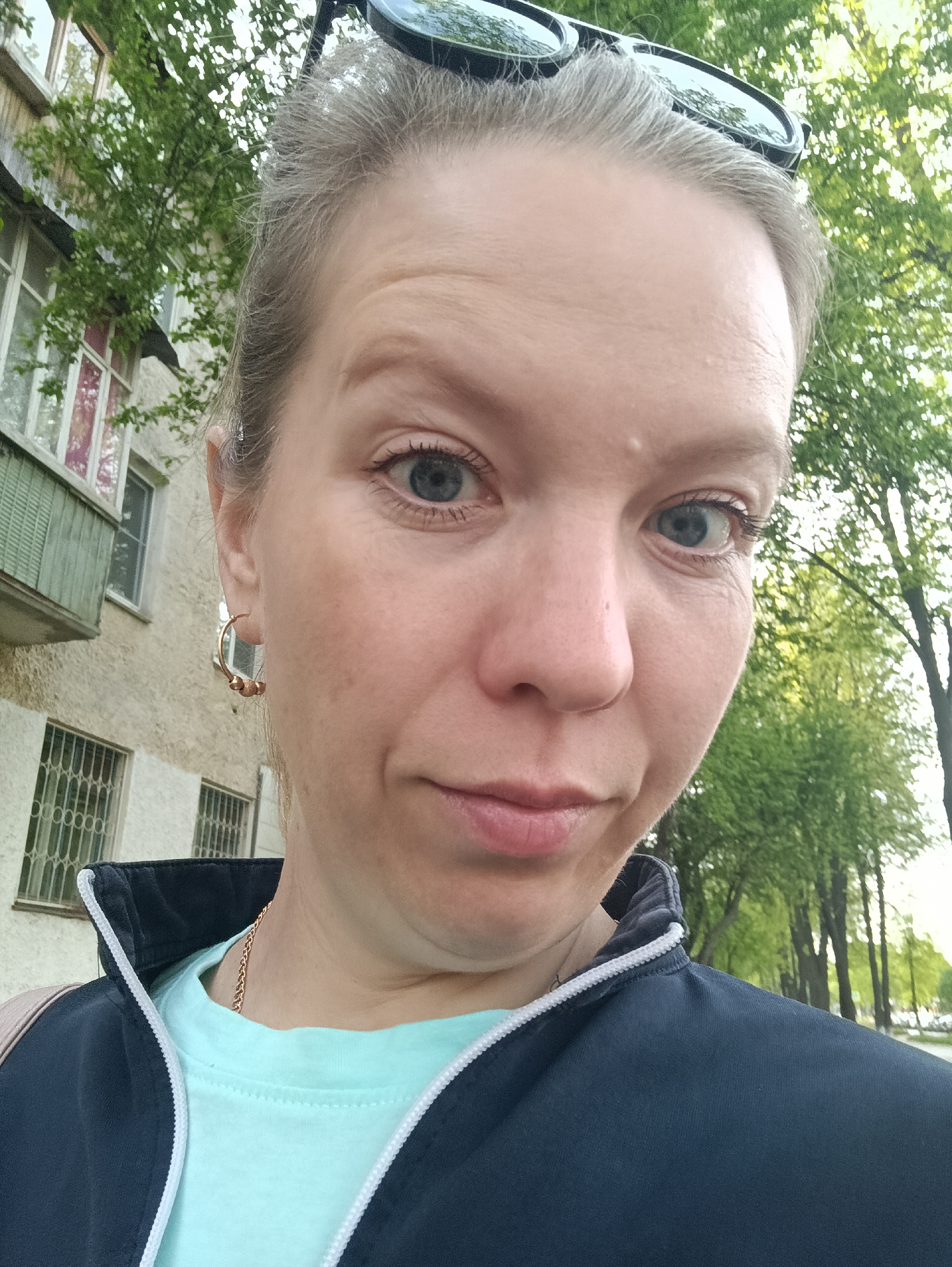 A close-up, selfie-style photograph depicts a woman with fair skin and graying blonde hair pulled back, wearing a light blue T-shirt and a navy jacket with white trim, posing outdoors. Sunglasses rest on top of her head. She has blue eyes and is wearing small, gold-colored hoop earrings. Behind her, a faded, multi-story building with barred windows and a balcony is visible. Trees with green foliage are present in the background. The overall impression is of a casual, daytime snapshot taken in an urban environment, likely in Chaikovsky, Russia, based on the user's prompt.