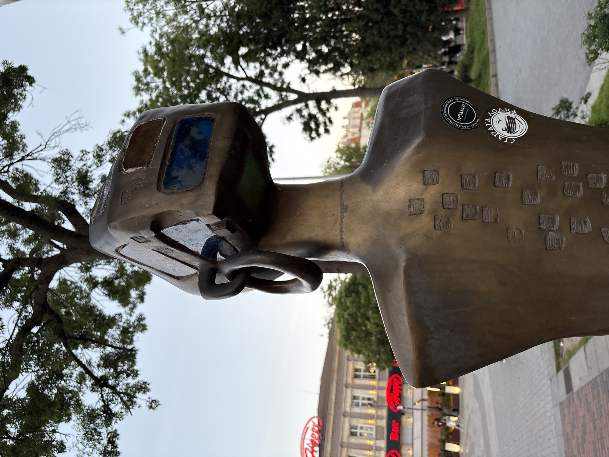 The image shows a close-up, low-angle view of a bronze sculpture in Varna, Bulgaria. The sculpture features a stylized head with boxy features and blue panels resembling windows. A pair of linked rings or loops encircle the neck area. Two stickers are affixed to the side of the head, one featuring the word "NOMAD" and the phrase "IN FOOD WE TRUST," and the other featuring Cyrillic text including the word "ЛОДКА" (Lodka/Boat) and what appears to be the word "СТАРА" (Stara/Old).
In the background, trees are visible, along with a section of a building that displays the "Happy Bar & Grill" logo. The ground appears to be paved. The image is slightly tilted, giving the impression that the sculpture is leaning.