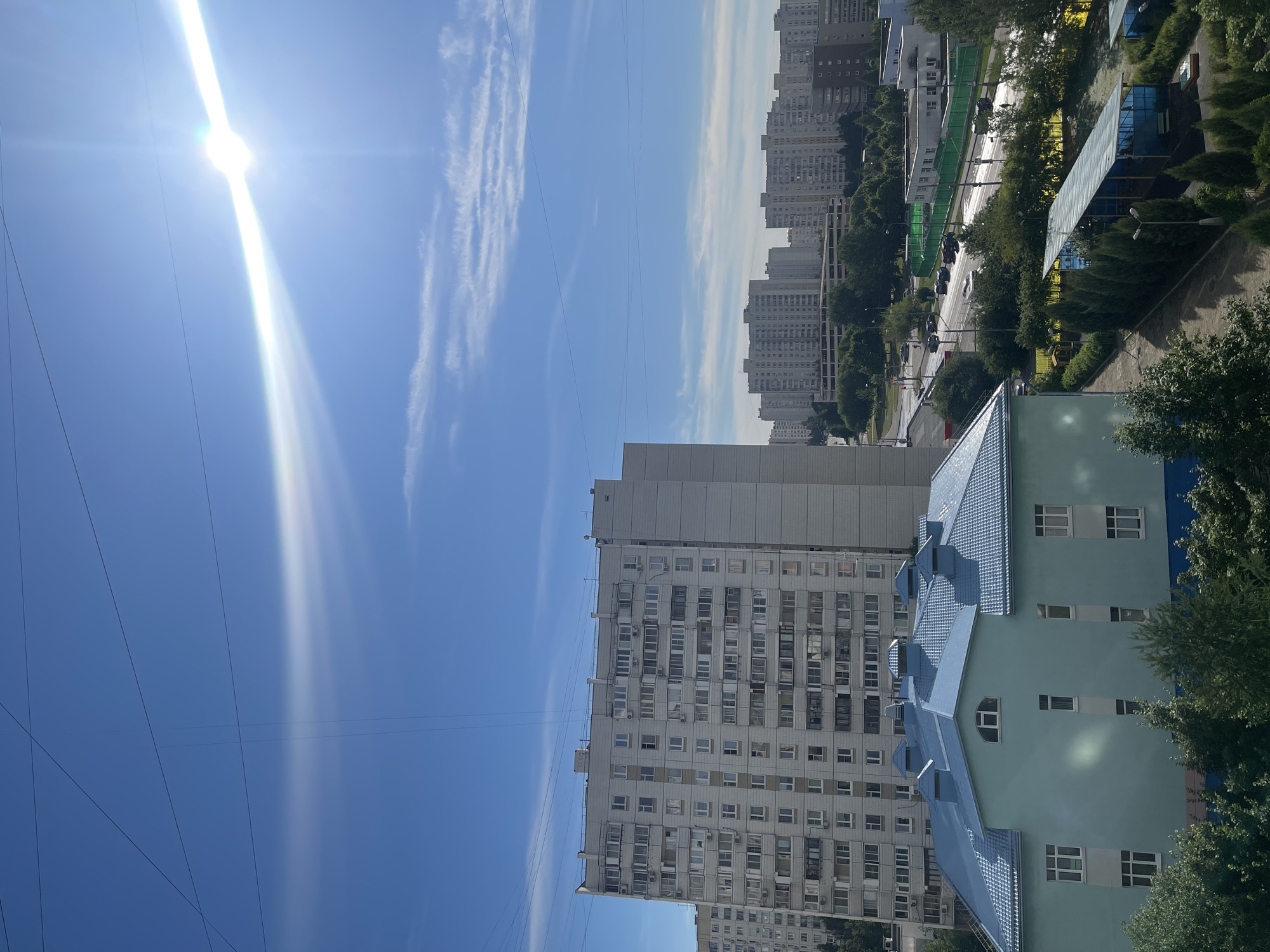 This is an outdoor, eye-level, medium shot of a residential area in Moscow, Russia. In the foreground, a light green building with a blue, multi-peaked roof is visible, with trees surrounding its base. Behind this, a tall, gray apartment building dominates the mid-ground, with numerous balconies and windows. Further in the distance, a dense cluster of similar apartment buildings stretches across the horizon under a bright blue sky with scattered wispy clouds and a strong sun glare. A road with passing cars and a green fenced area are visible between the apartment buildings and the foreground structures. Overhead, several power lines crisscross the sky.
