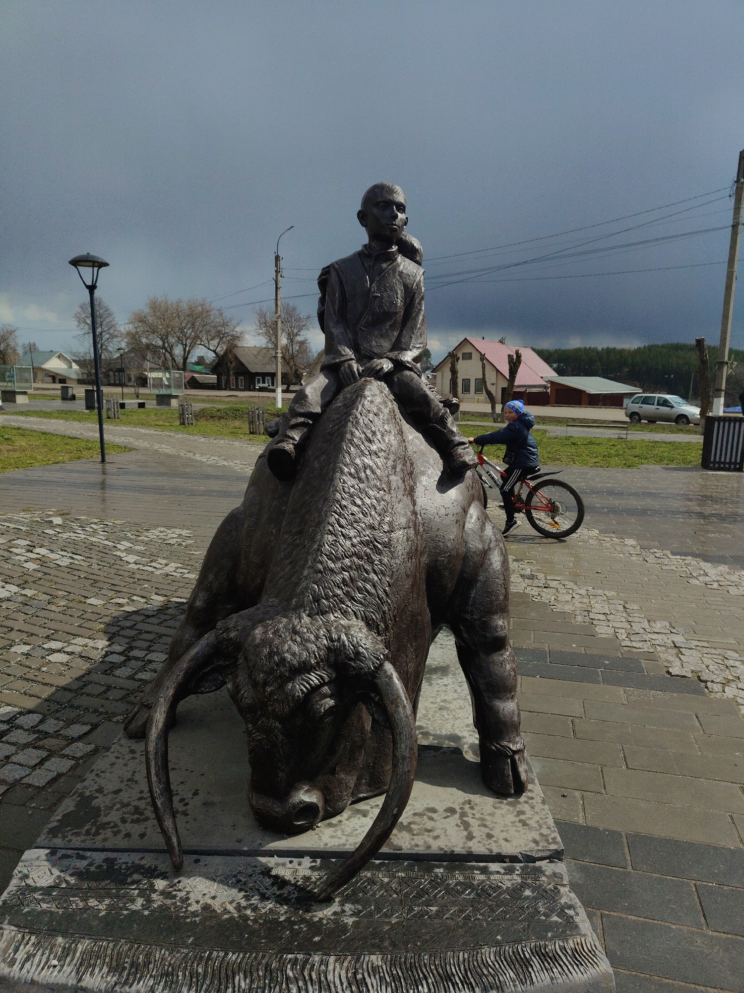 The image depicts an outdoor scene dominated by a bronze statue of a person riding a bull. The statue features a figure, possibly a youth or young man, seated atop a large, textured bull that appears to be resting or kneeling. The bull has prominent horns and a rugged, sculpted hide. The base of the statue is patterned, resembling a decorative rug or blanket, and shows signs of being wet.
In the mid-ground, a child wearing a blue hat and jacket is riding a red and white bicycle on a paved path. The background consists of several houses, some with red roofs, and bare trees, suggesting a village or small town setting. Utility poles with overhead wires and streetlights are also visible. Further back, a white car is parked.
The sky is largely overcast with dark, heavy clouds, indicating recent rain or impending stormy weather, though some brighter areas suggest the weather might be clearing. The ground beneath the statue and along the paths is wet, confirming the presence of moisture. The pathways are made of a combination of cobblestones and rectangular pavers. No discernible text is visible in the image.