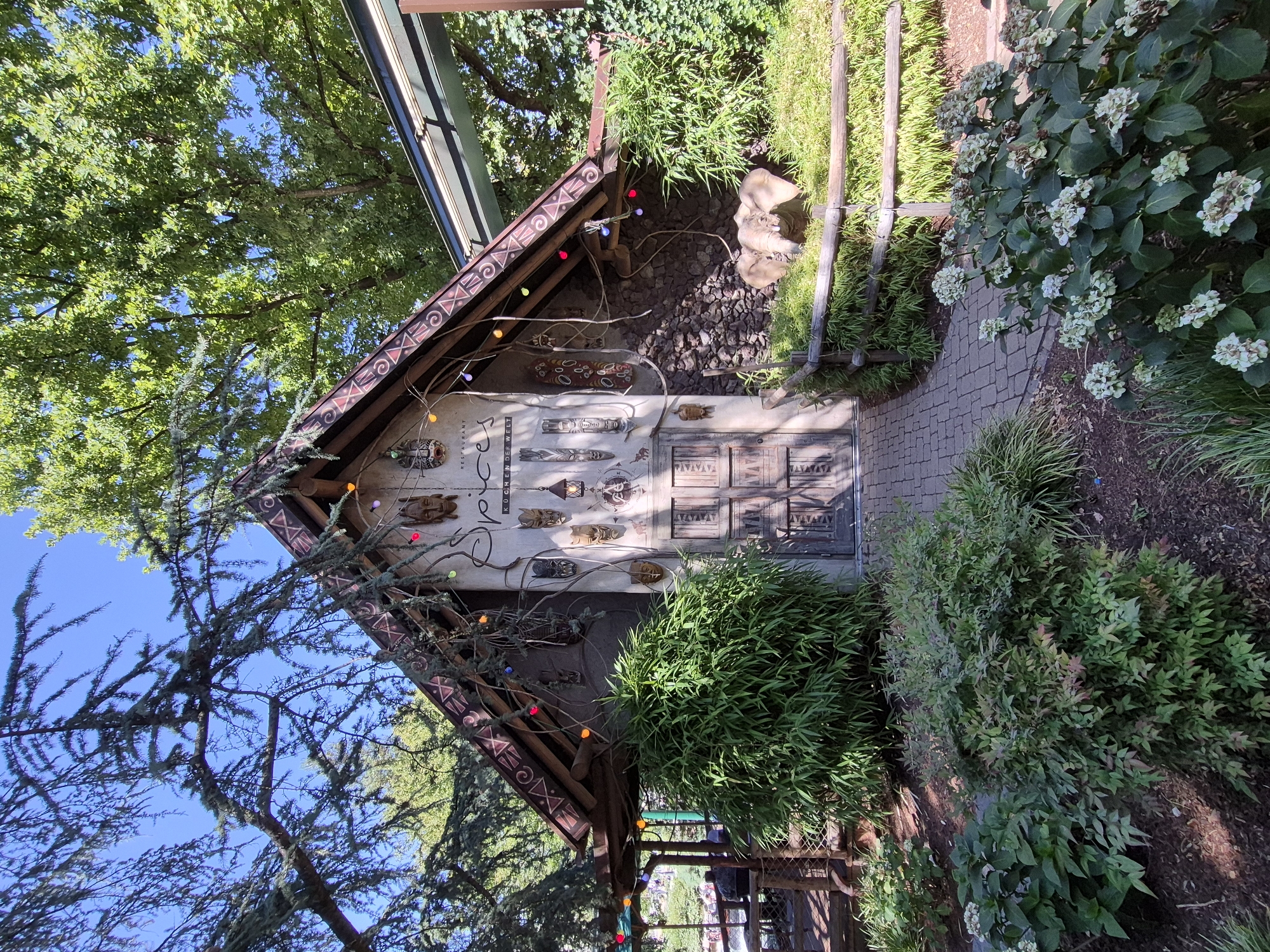The image shows the entrance to a restaurant called "Spice" in the city of Rust, Germany. The entrance is decorated with various tribal masks and colorful lights. There are plants and trees surrounding the entrance, and a small elephant statue is visible in the garden. The overall atmosphere is exotic and inviting.