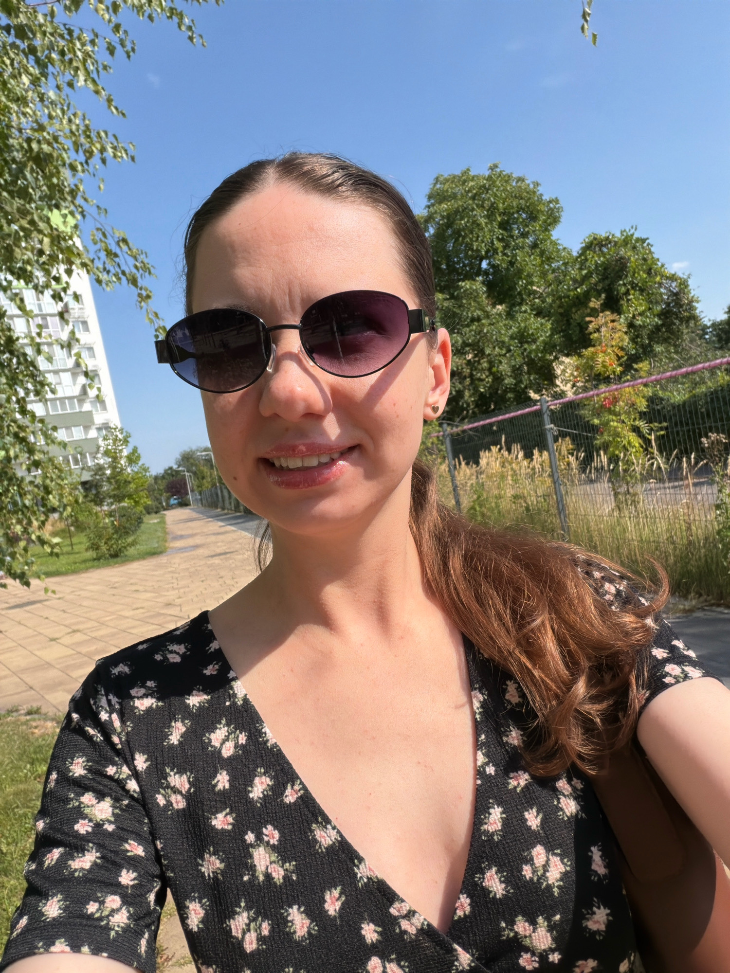 This is a selfie of a woman with brown hair pulled back in a ponytail, wearing dark-rimmed sunglasses and a black floral dress. She is smiling and standing outdoors on a sunny day. Behind her, a paved pathway is visible, bordered by grass and trees. To the left, a tall, modern apartment building stands against a clear blue sky. To the right, a chain-link fence with tall, dry grass behind it is present. The scene appears to be a park or residential area within the city of Kyiv, Ukraine.