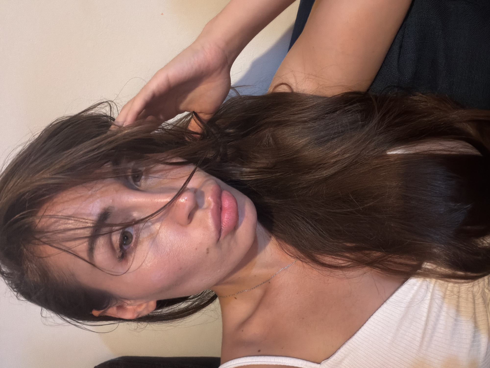 A woman is posing for a selfie in a dimly lit room. She has long, dark brown hair that is partially covering her face. Her eyes are brown, and she is wearing a white tank top. Her left hand is resting on her hair, and her right arm is visible in the background. The background is a plain, light-colored wall. The image appears to have been taken indoors, likely in a residential setting. There is no specific identifiable information within the image to determine its location in the city of Tersefanou, Cyprus.