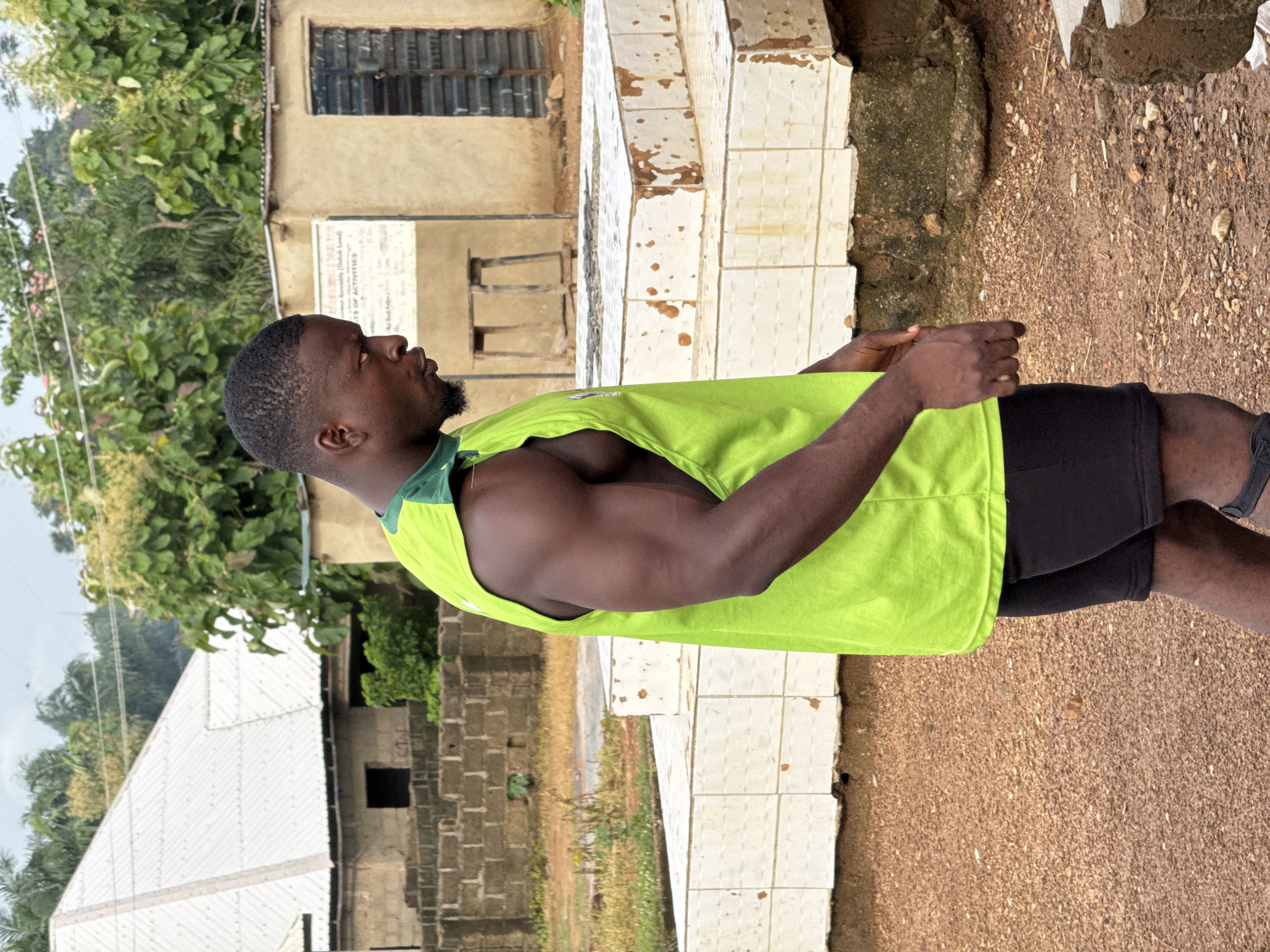 A young Black man wearing a neon green sleeveless athletic shirt and black shorts stands in an outdoor area, facing right and looking down. His musculature is prominent, particularly in his arms and shoulders. The background features a low-slung building with a tan facade and dark-framed windows, alongside a white tiled structure that appears to be a monument or well. Beyond that, a building with a corrugated metal roof and brick walls is visible, surrounded by sparse vegetation and trees under a cloudy sky. The ground is covered in reddish-brown soil and gravel. The image appears to have been taken in Akwanga, Nigeria, judging by the architectural style and natural elements suggestive of the region.