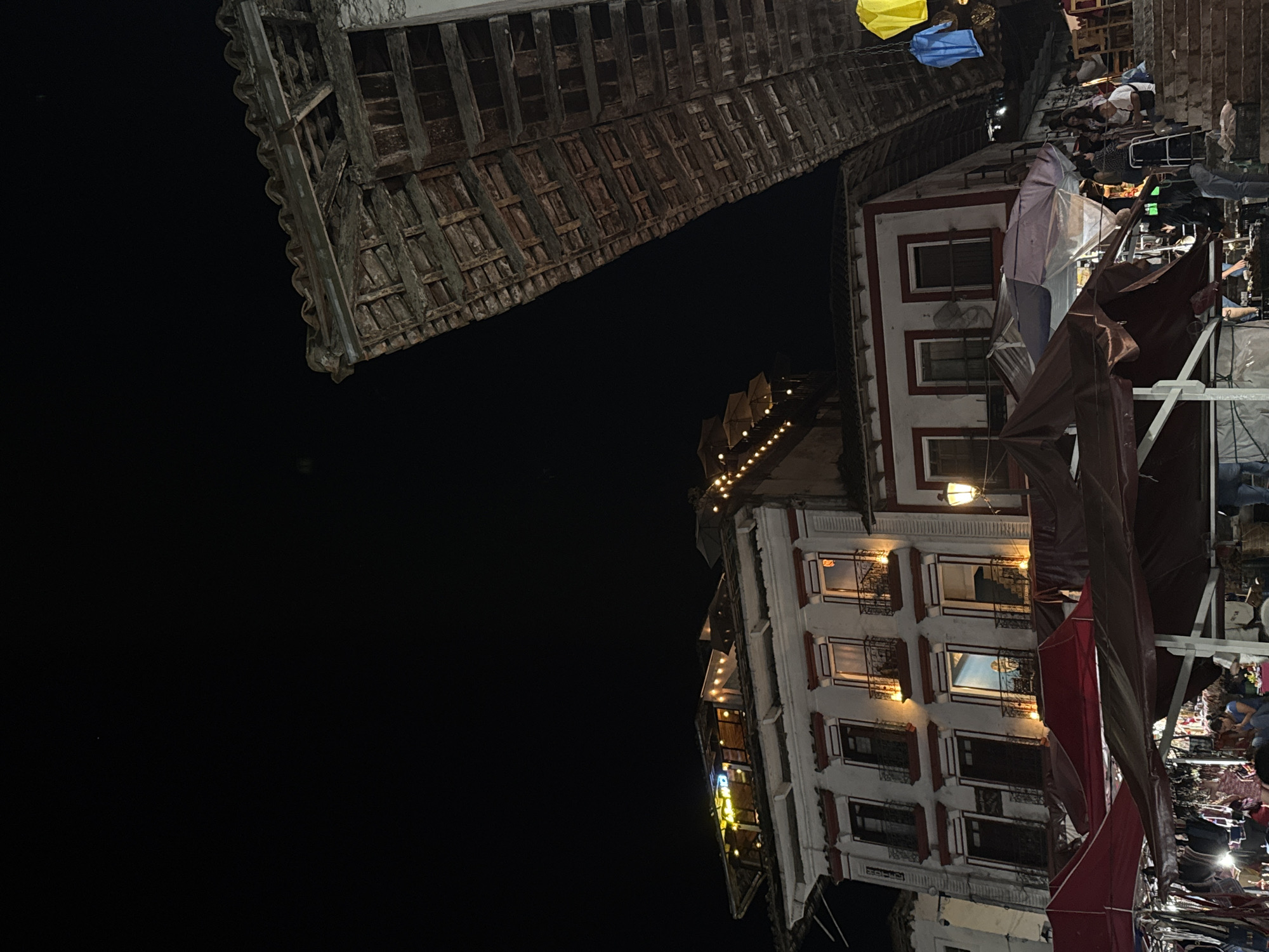 The image shows a view of the night market in Cuetzalan, Mexico. The foreground contains the dark night sky, taking up over half of the frame. In the middle ground, there is a structure with old, wooden latticework. Adjacent to that, there is a multi-story building with a white facade and dark window frames. String lights and lanterns illuminate the building. In the lower right portion of the frame, there are market stalls covered with red and brown tarps. People are present, but not easily discernible.