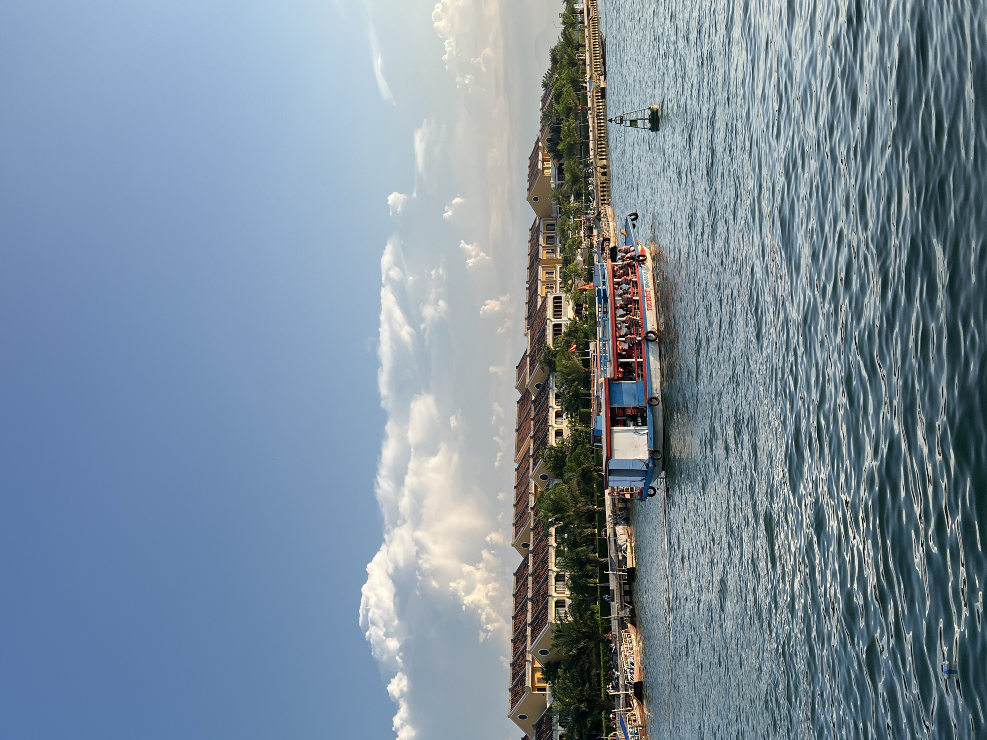 The image shows a sunny day scene in Hội An Ward, Vietnam. A red, white, and blue tourist boat, filled with passengers, is docked alongside a waterfront lined with multi-story buildings, likely hotels or residences. A navigation buoy is visible in the water near the boat. The sky is mostly clear blue with scattered white clouds. The water is slightly choppy, reflecting the sunlight. Palm trees are interspersed amongst the buildings. Other boats are docked nearby.