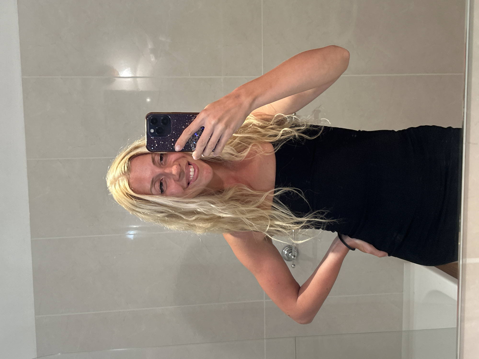 A woman with long, wavy blonde hair takes a selfie in a bathroom. She is wearing a black strapless dress and has her left hand on her hip. Her right hand holds a dark-colored phone with a starry pattern. The bathroom has light-colored tiled walls and a reflective surface, likely a mirror, in the foreground. There is a white bathtub visible below the tiled area. The lighting is bright, and there are no other people or significant objects in the frame. The location is identified as Polincove, France.